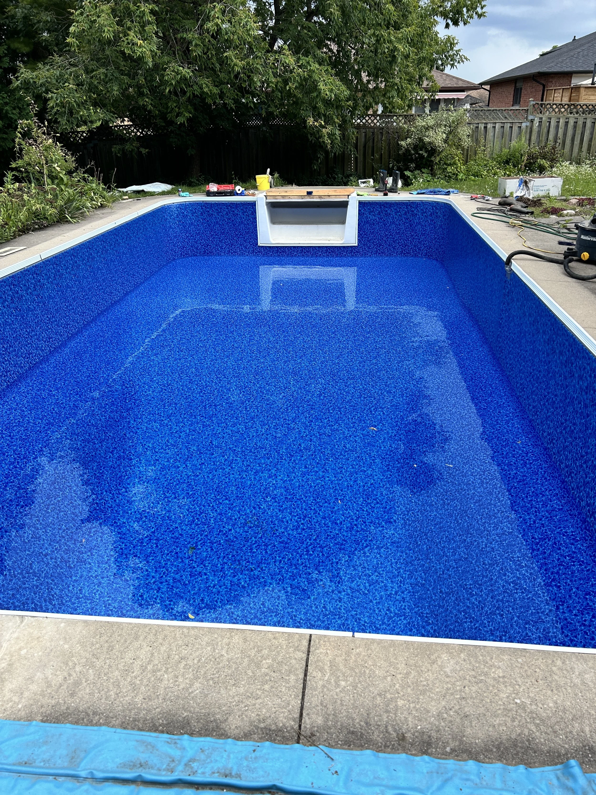 In-ground swimming pool with a bright blue interior, under construction, with tools and equipment around the edge in a backyard.