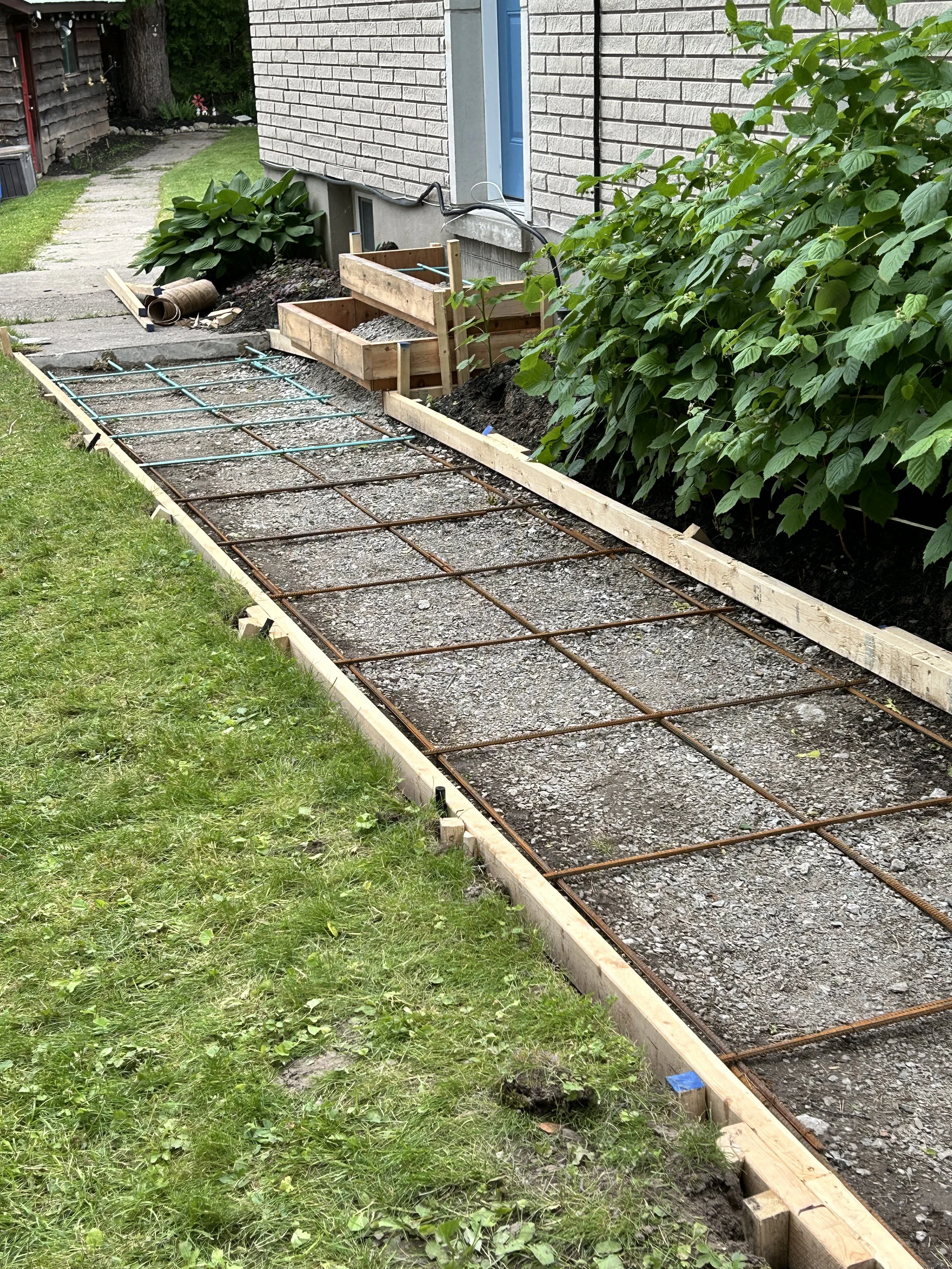Under construction pathway with wooden frame and metal rebar laid out, beside a house and green plants.
