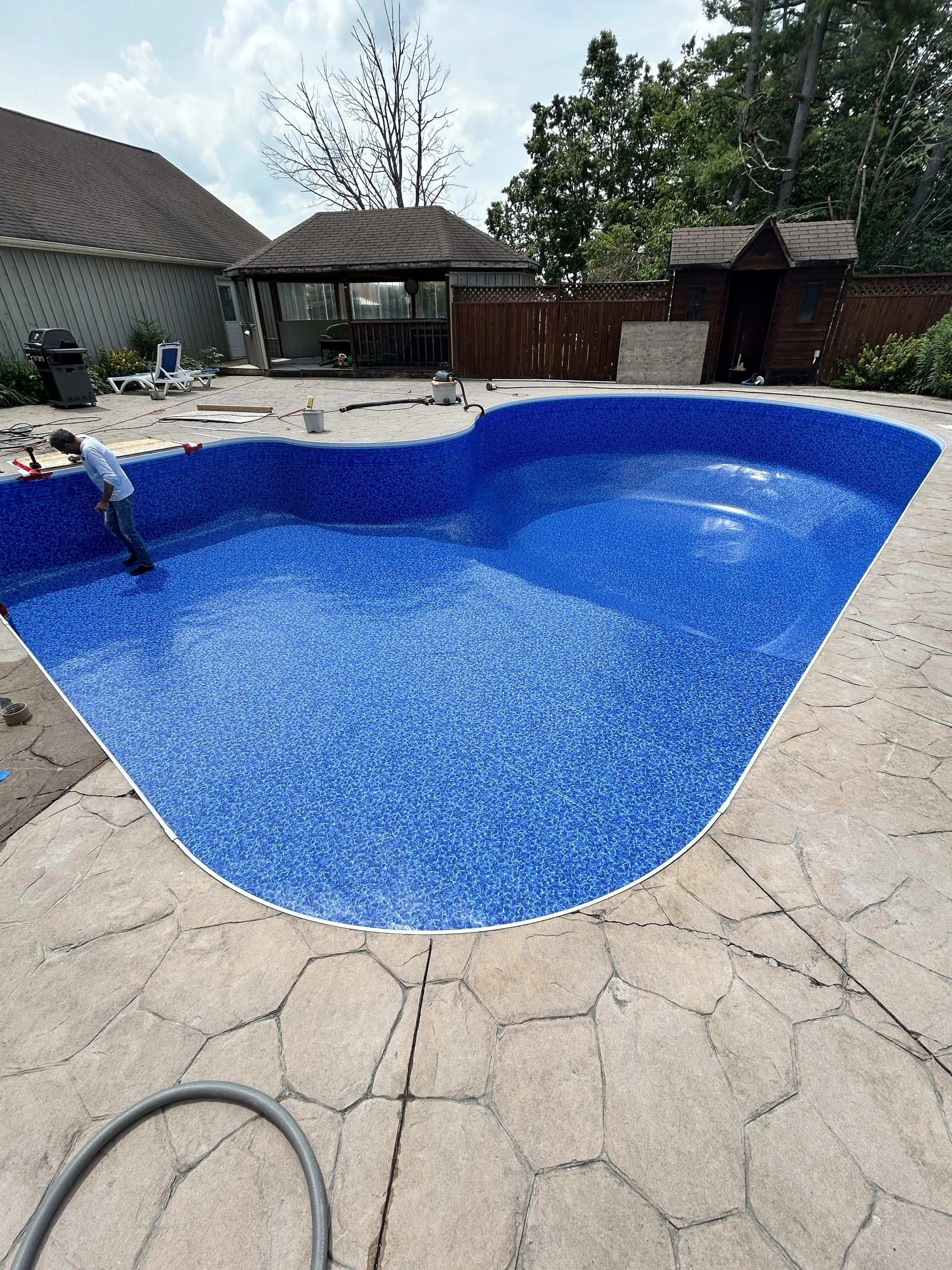 A backyard with an in-ground swimming pool under maintenance, with a person inside working on it. The area around the pool is paved with large stone tiles, and there are trees and a wooden shed in the background.