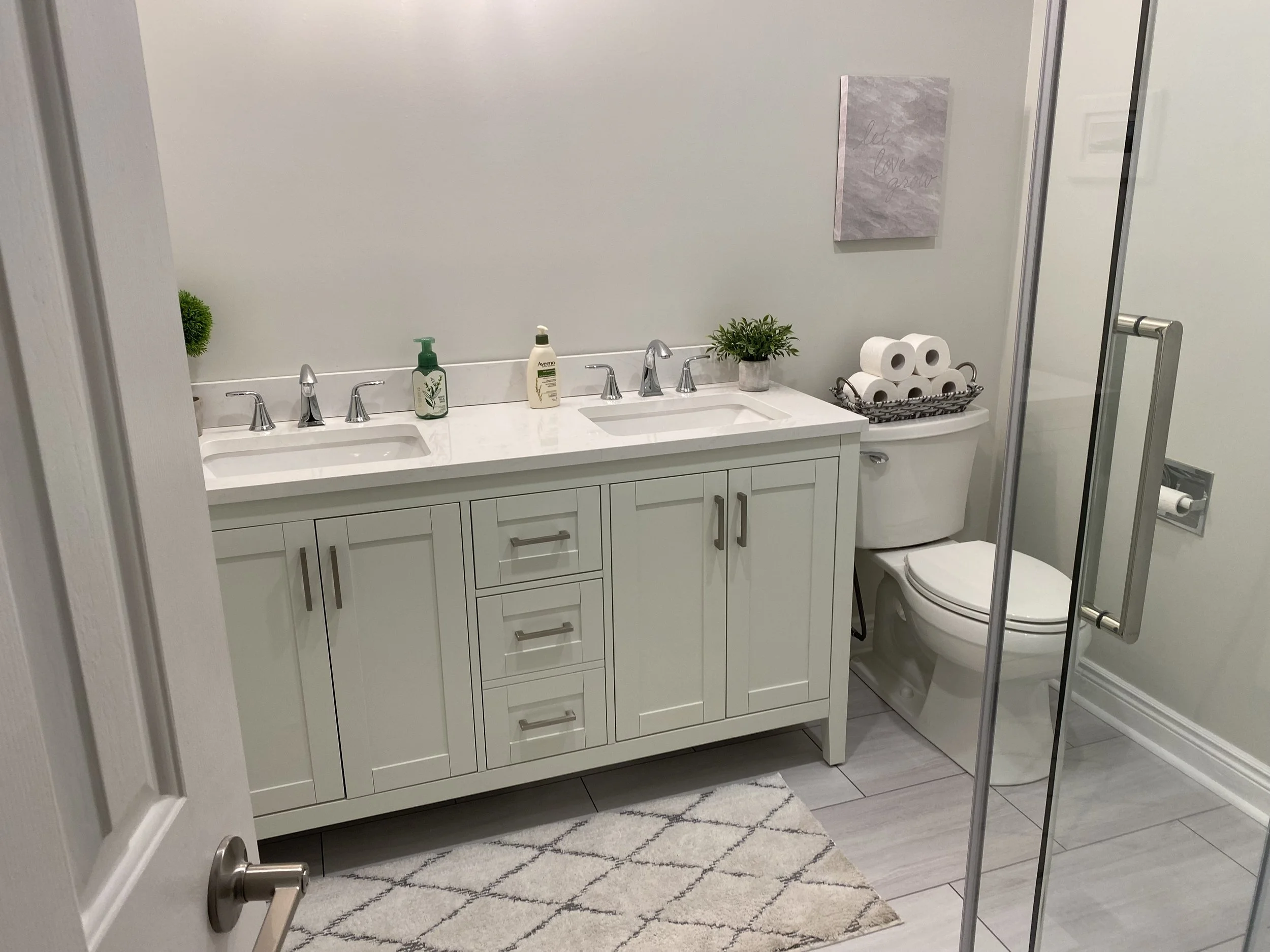A modern bathroom featuring a white vanity with two sinks, a framed mirror, and decoration including small plants, soap dispensers, and a basket of toilet paper rolls. A toilet is visible next to a glass shower door, with a framed wall art piece abov