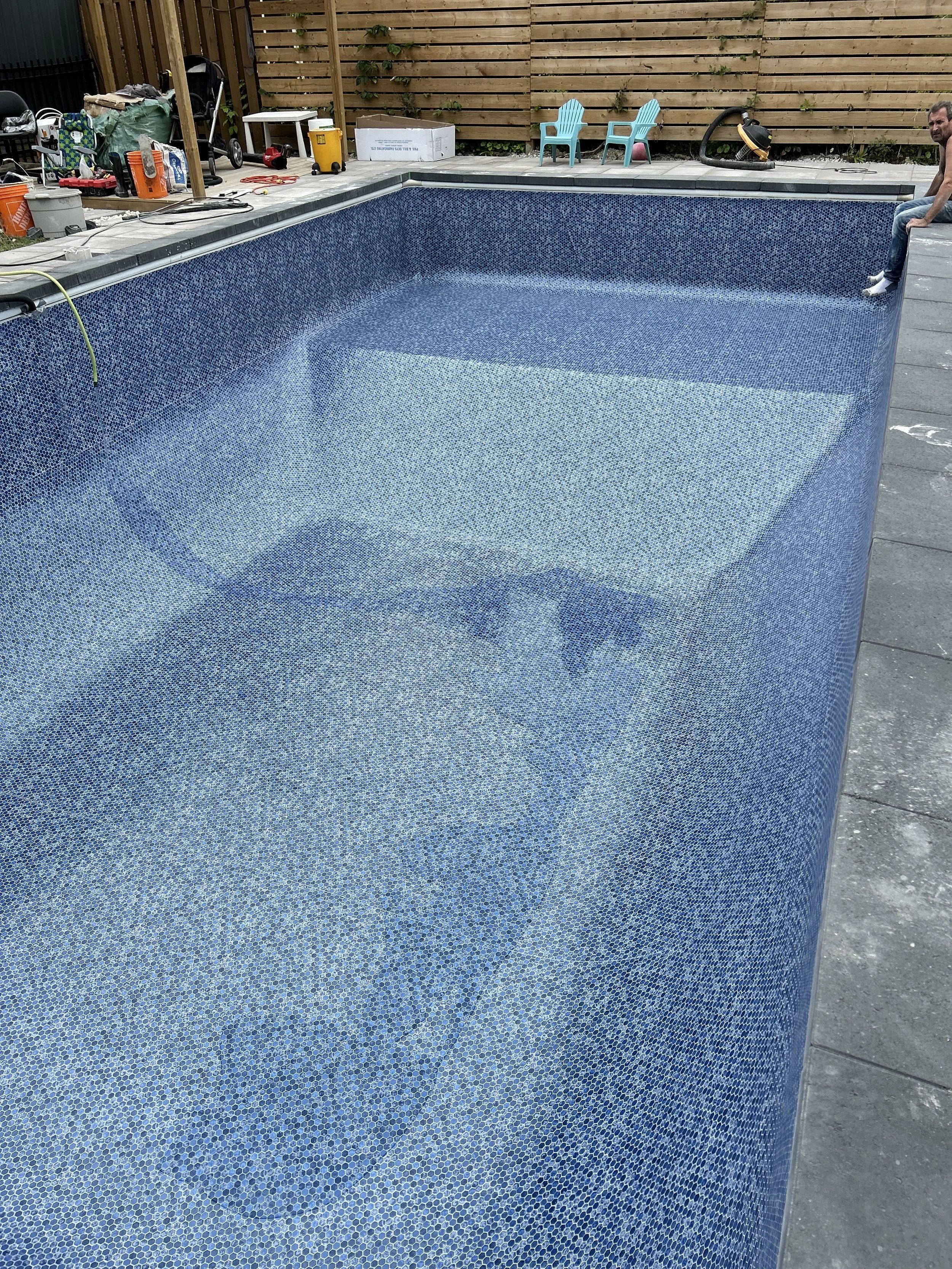 A swimming pool under renovation with mosaic tiles inside, partially empty and surrounded by a deck with various tools and furniture.