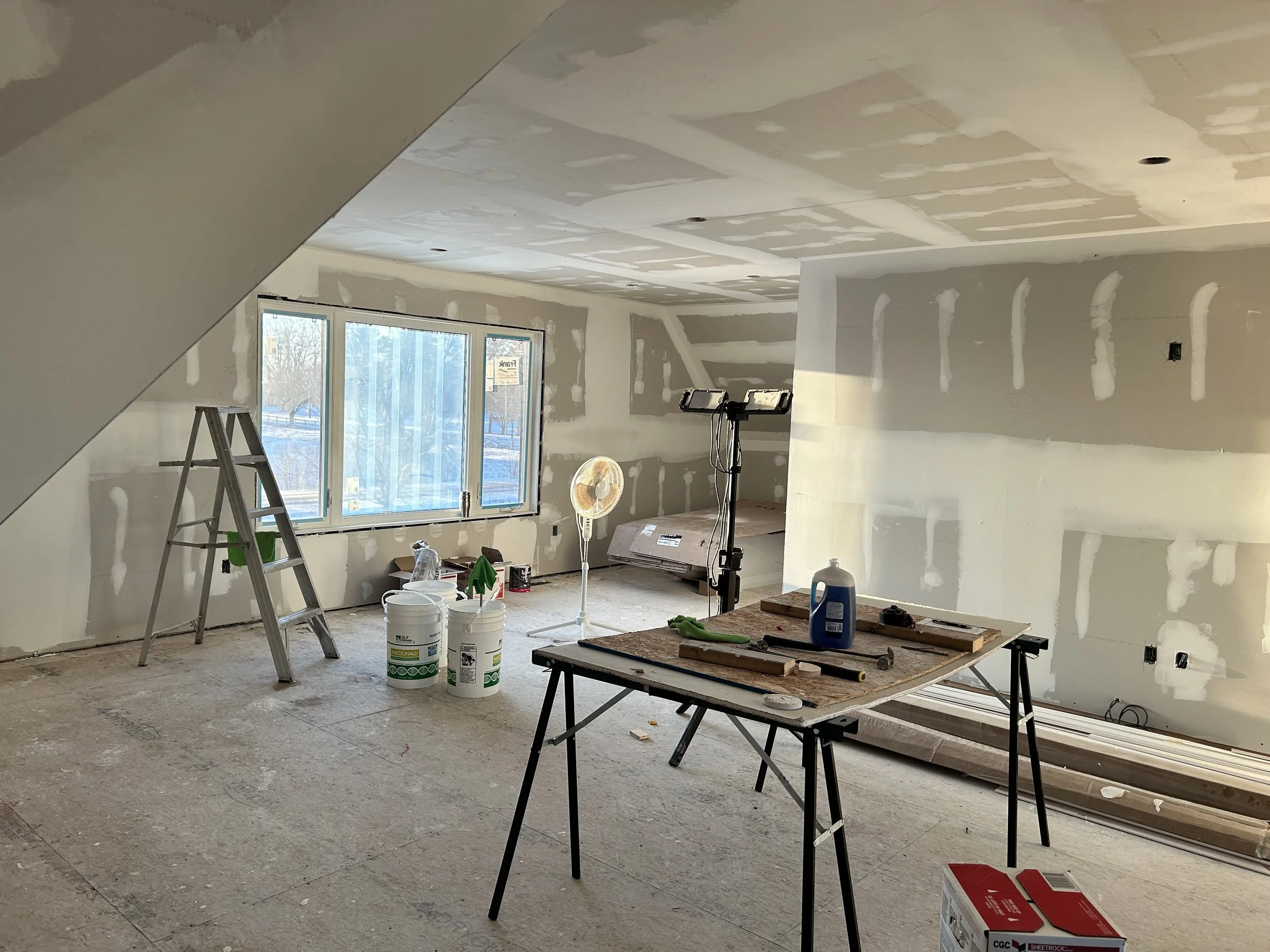Room under construction with drywall walls, a ladder, buckets of paint, a standing fan, tools on a work table, and large windows.