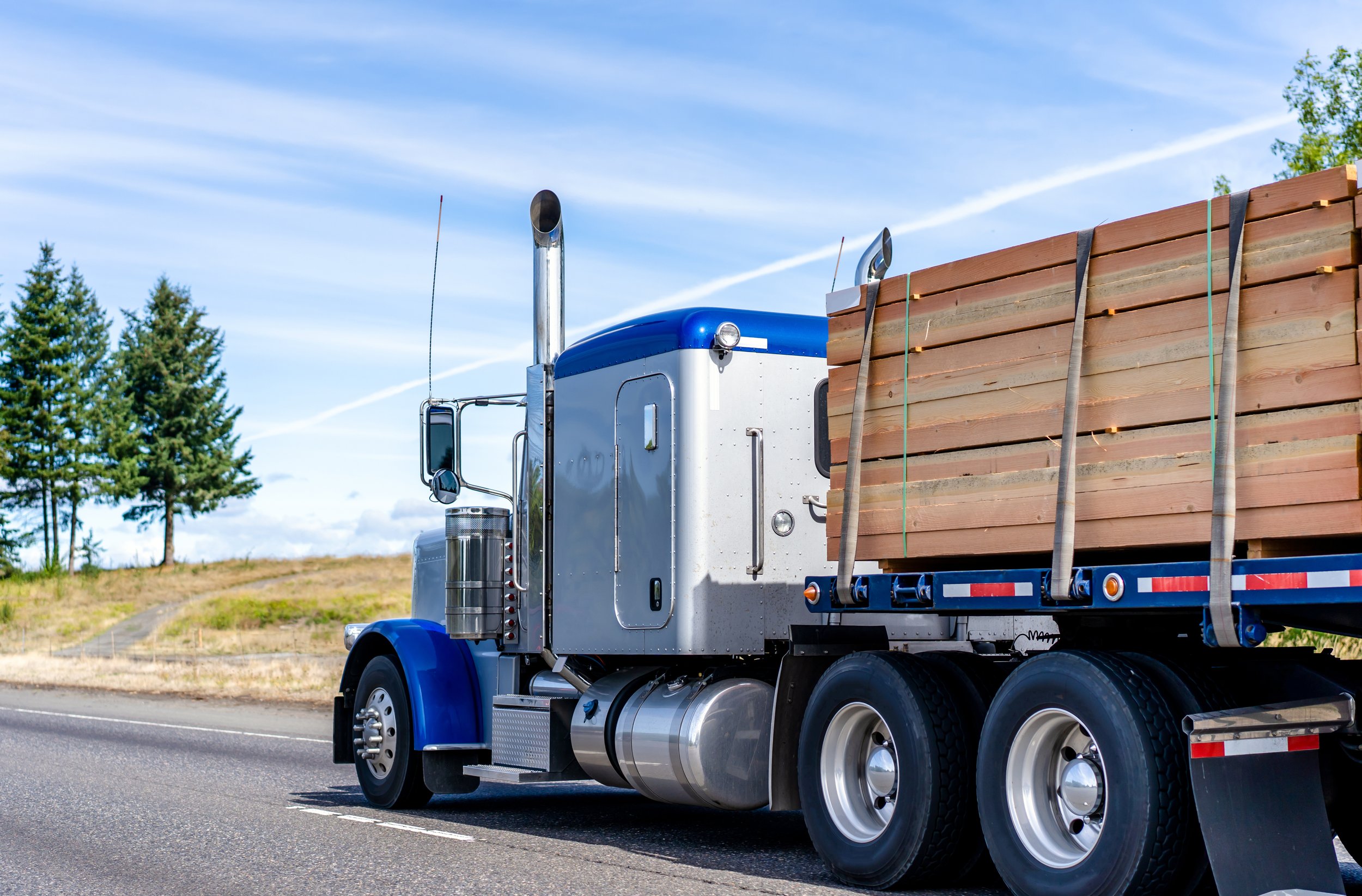 Blue and gray semi-truck transporting wooden beams on a highway with trees and a clear sky in the background.
