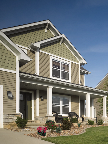 Suburban house with front porch, green siding, and white trim surrounded by plants and flowers.