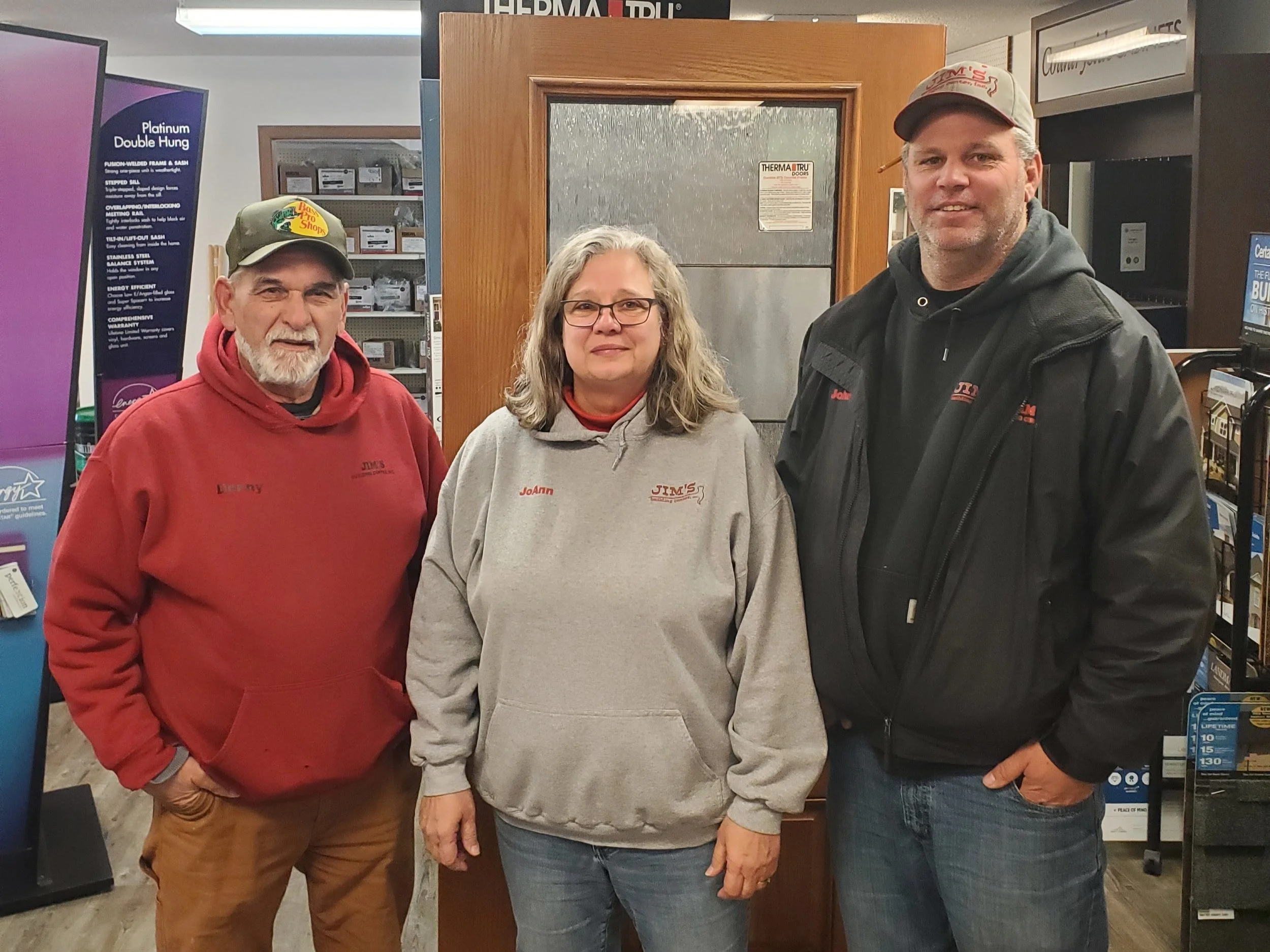 Three people posing in a showroom, wearing branded clothing and hats, standing in front of a door display.