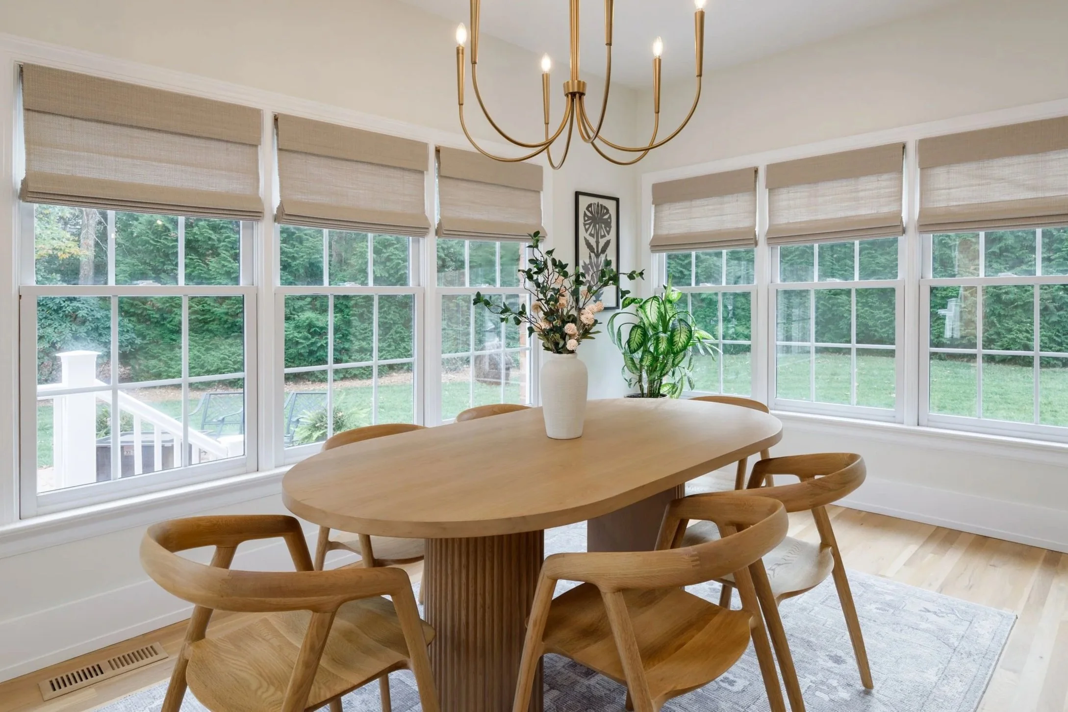 Bright dining room with large windows, beige Roman blinds, a wooden table with six matching chairs, a chandelier, green plants, and a framed flower art on the wall.