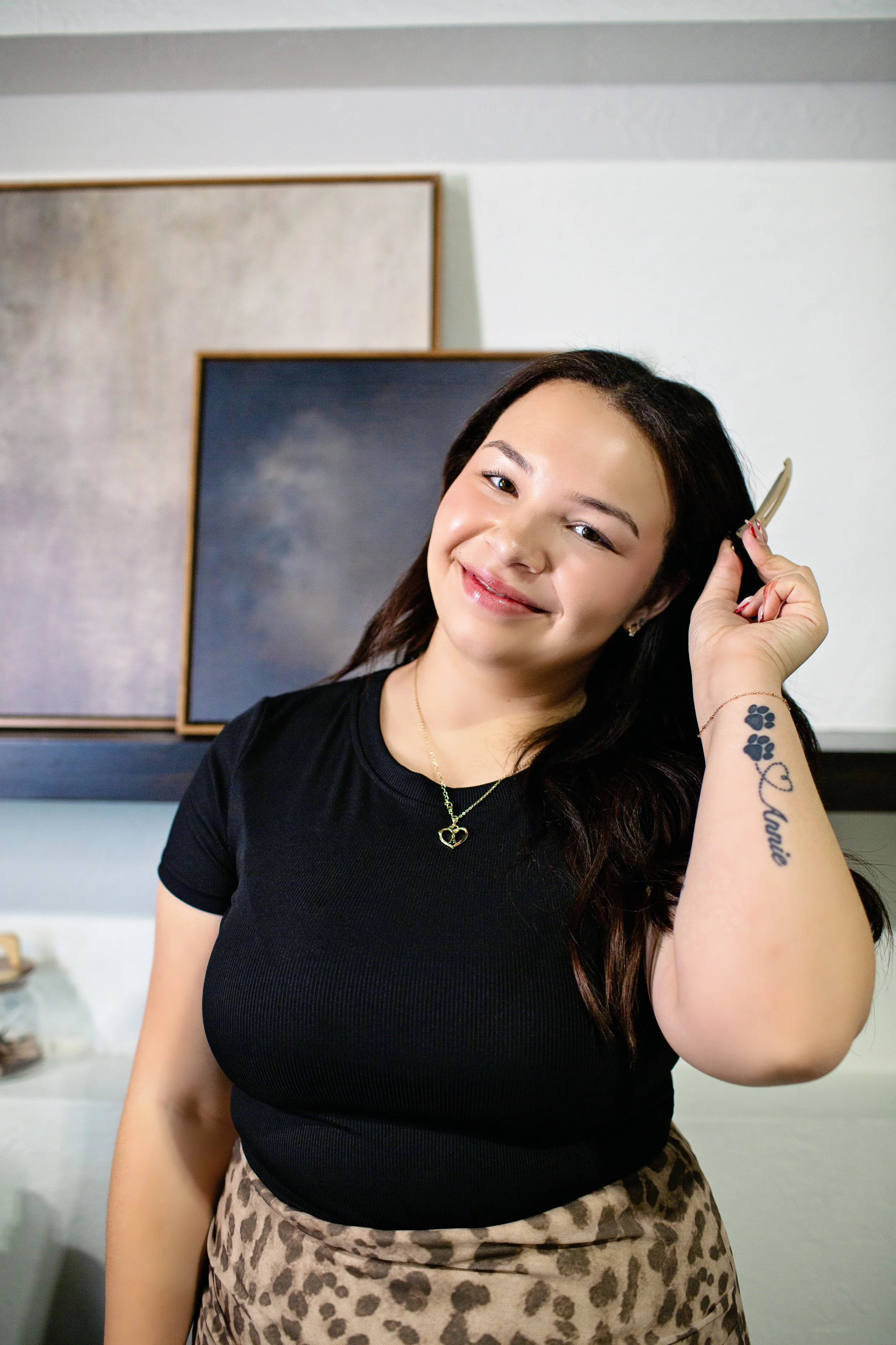 A smiling woman with dark hair is holding hair clips near her head in a room with abstract artwork on the wall behind her.