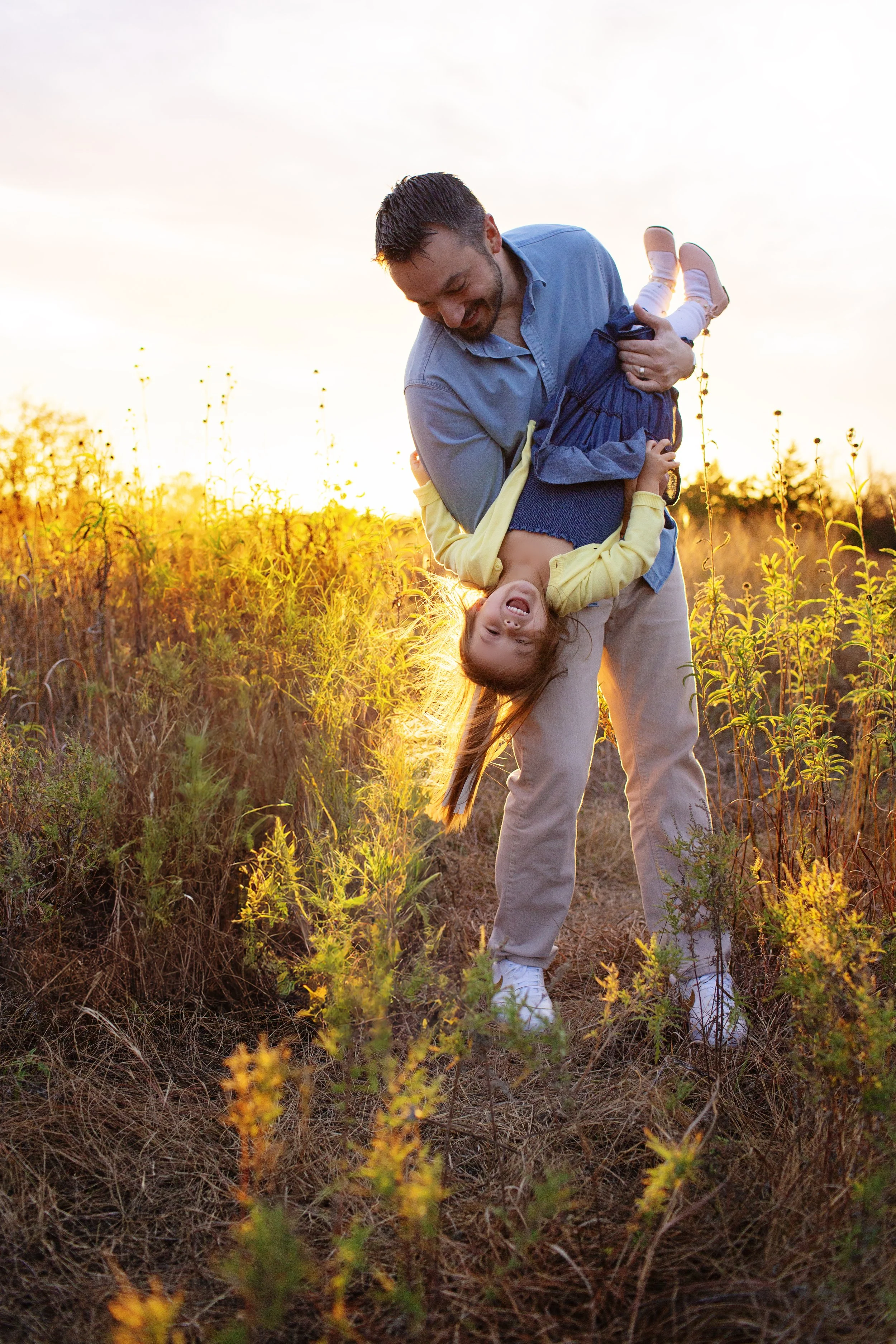 A man and young girl playing and having fun outdoors in a field with yellow wildflowers at sunset. The man is holding the girl upside down, and both are smiling and laughing.