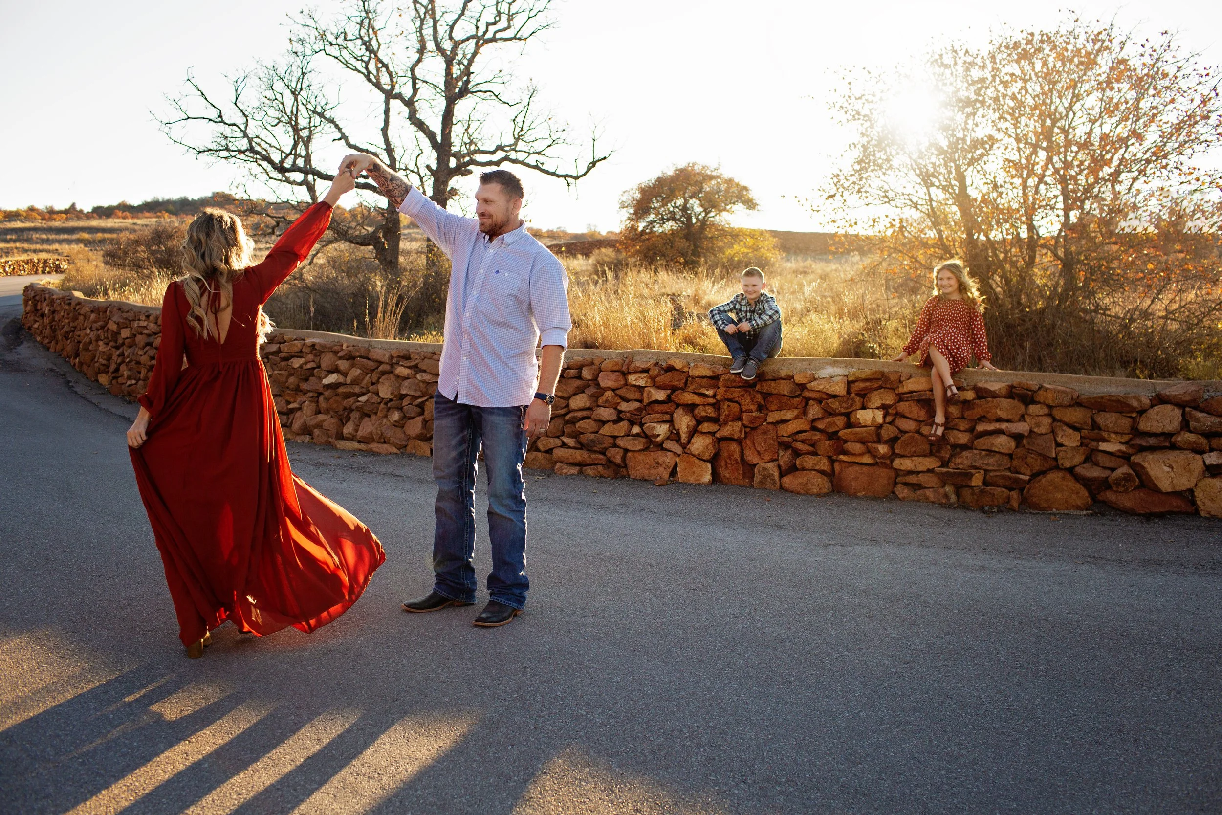 A couple dancing on a road in front of a stone wall with two children sitting on it, during sunset in a rural area with trees and open fields.