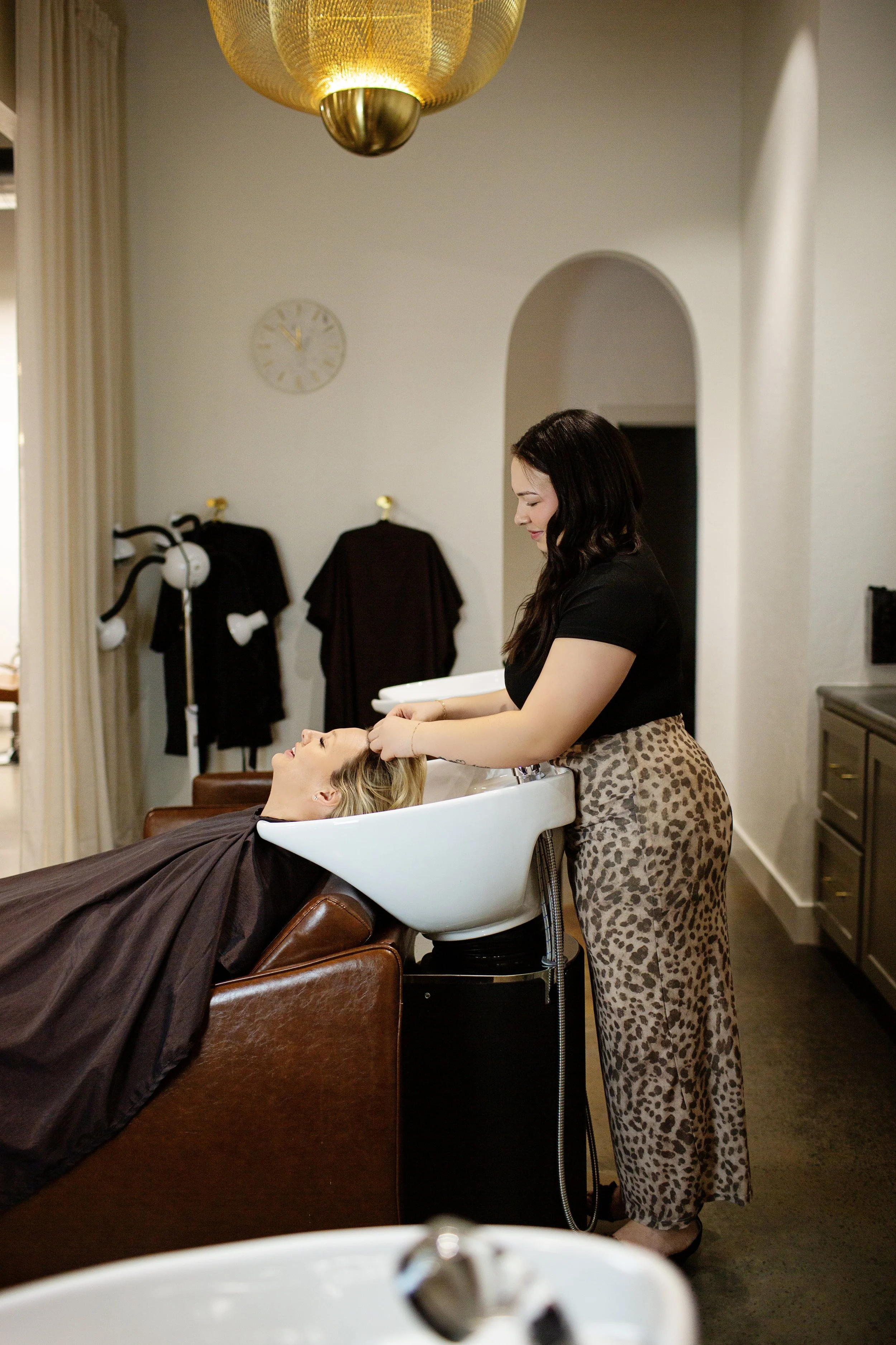 A woman with blonde hair is reclining in a salon chair with her head in a white shampoo basin, while a hairstylist with dark hair and leopard print pants washes her hair.