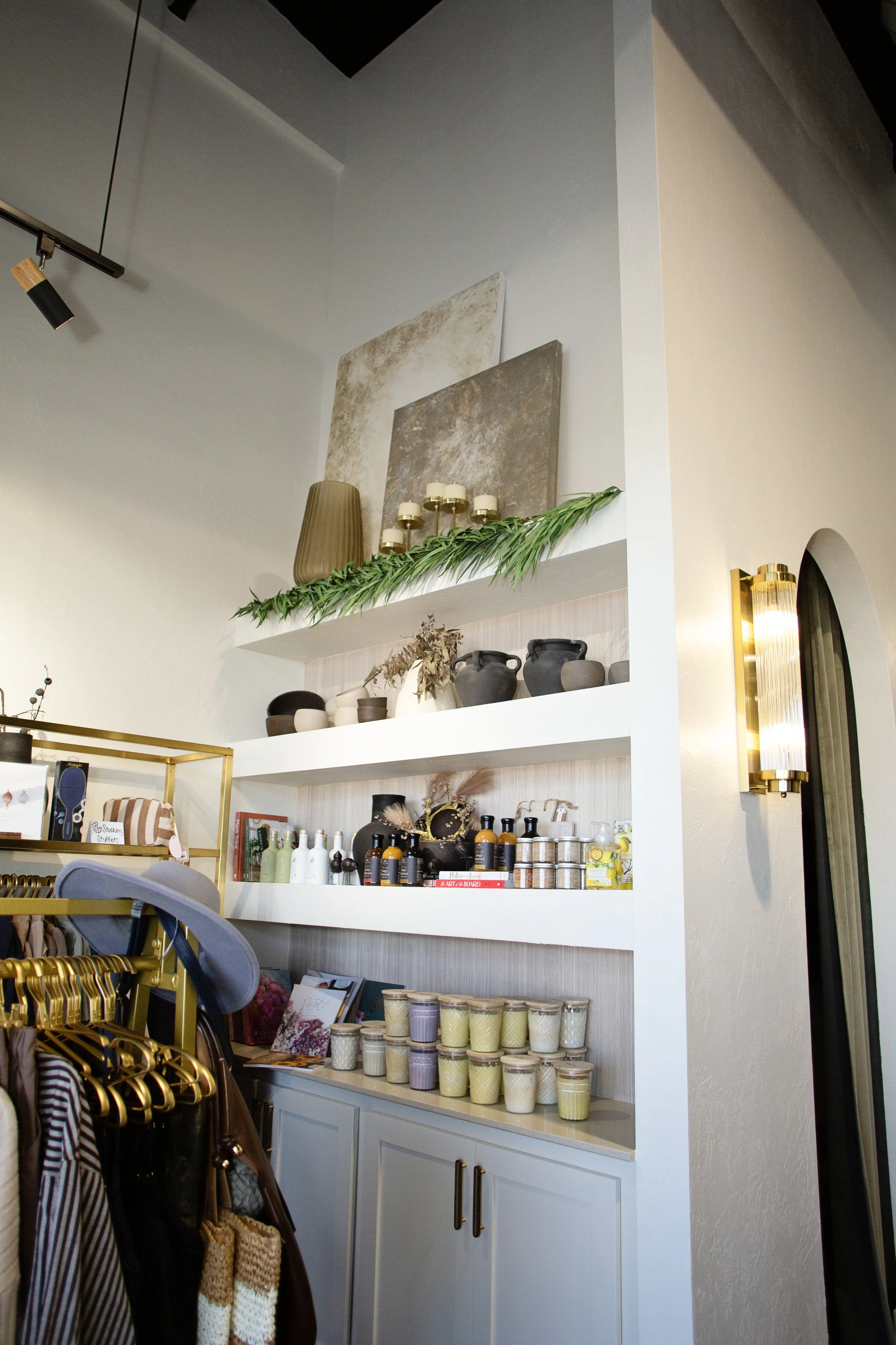Interior of a boutique or home decor store with white shelves displaying vases, candles, and decorative items. A clothing rack with hats and scarves is visible in the foreground. The wall features modern lighting fixtures and minimalist decor.
