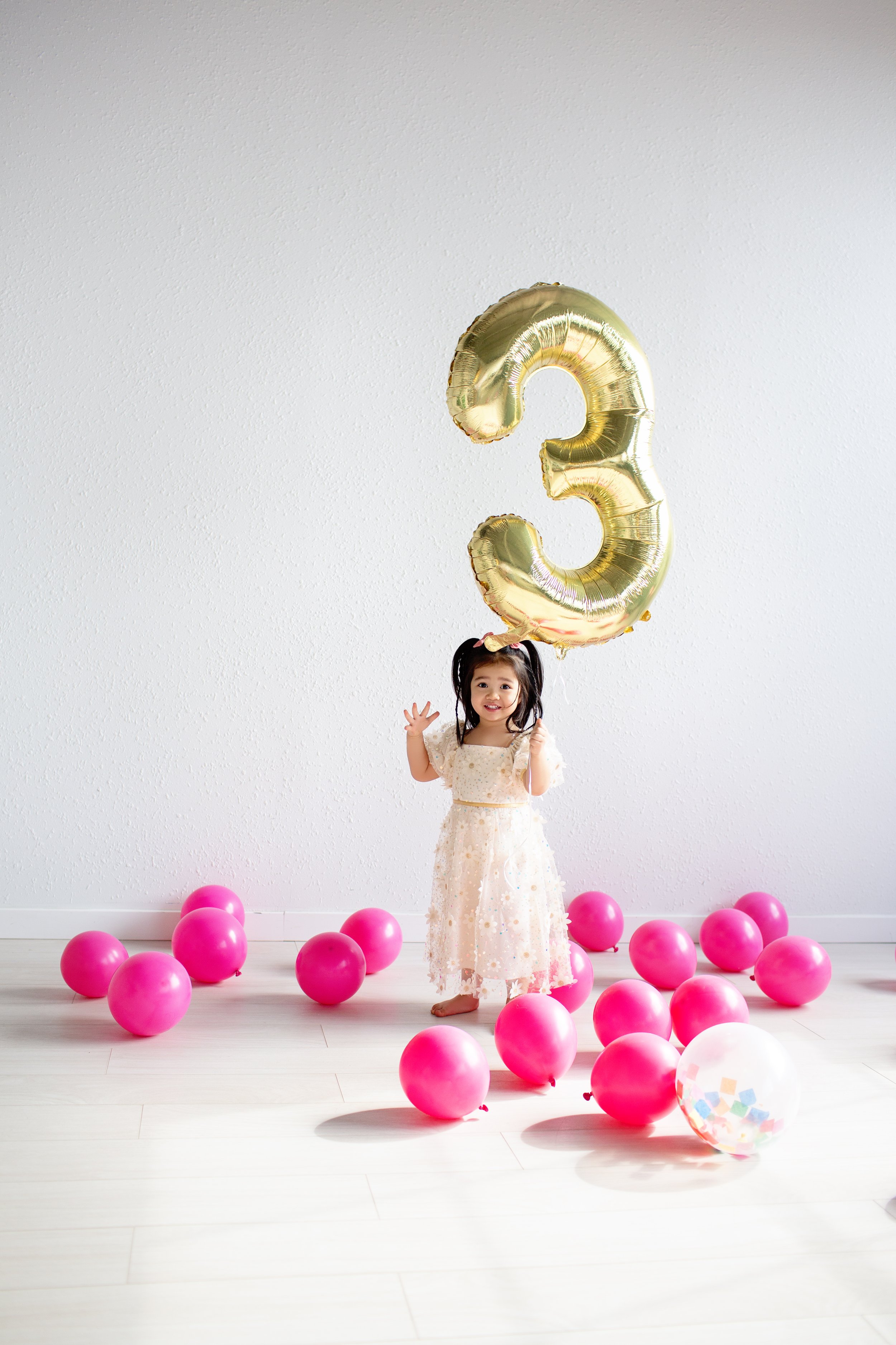 A young girl with dark hair in pigtails, wearing a light-colored dress, standing on a white floor surrounded by pink balloons, holding a large gold number three balloon, celebrating her third birthday.
