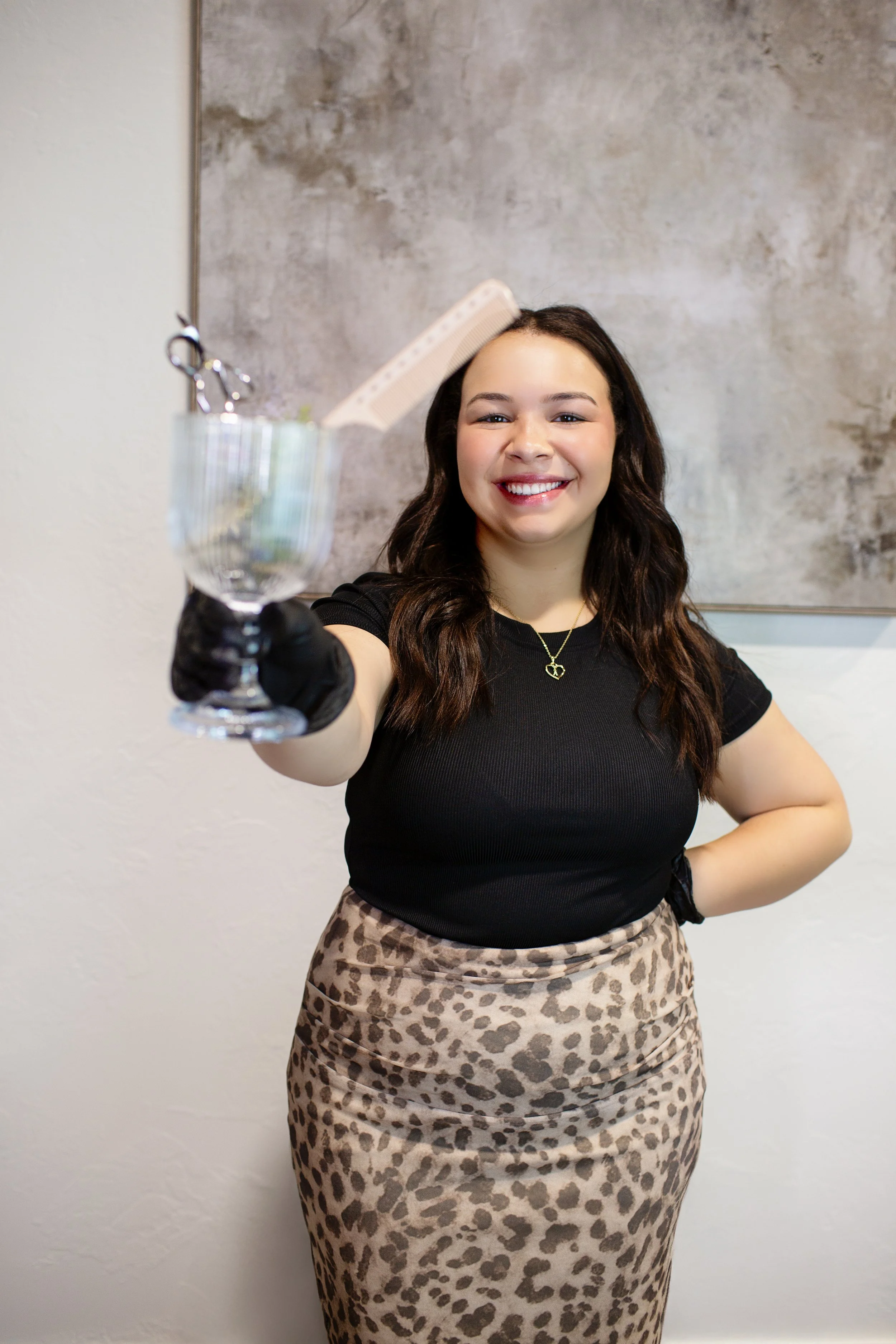A young woman with long dark hair smiling and wearing a black top and leopard print skirt, holding hairdressing scissors and a comb in a glass container.