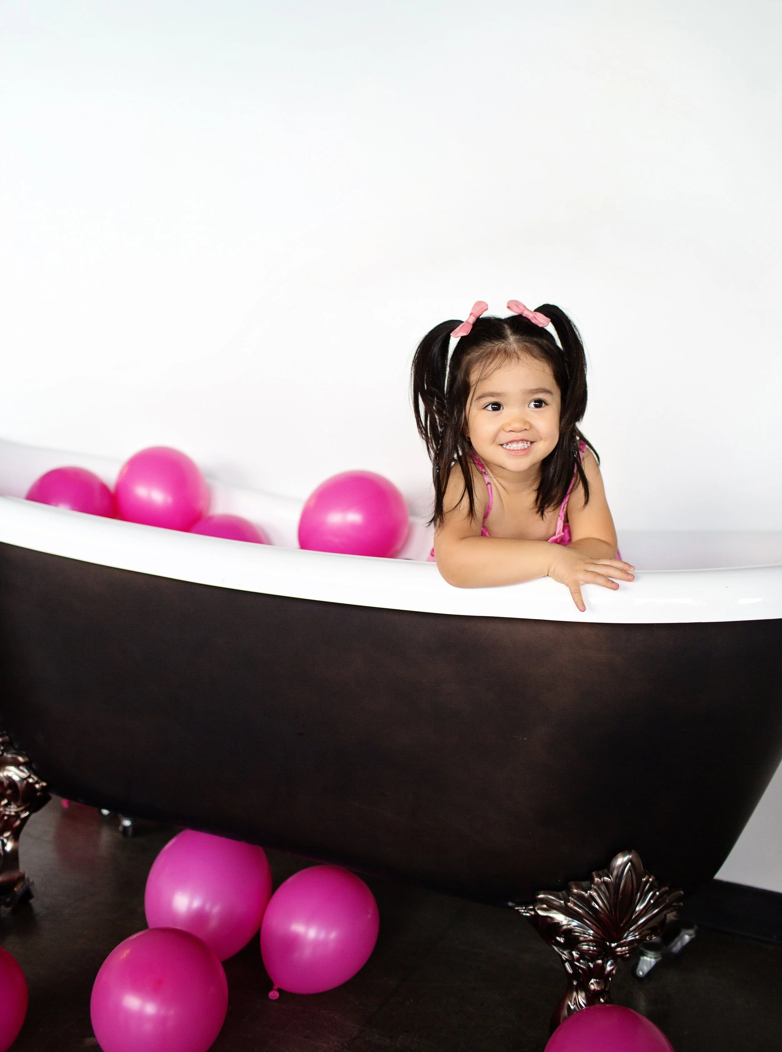 A young girl with pigtails and pink bows is smiling inside an abandoned black bathtub filled with pink balloons, with more pink balloons scattered on the floor.