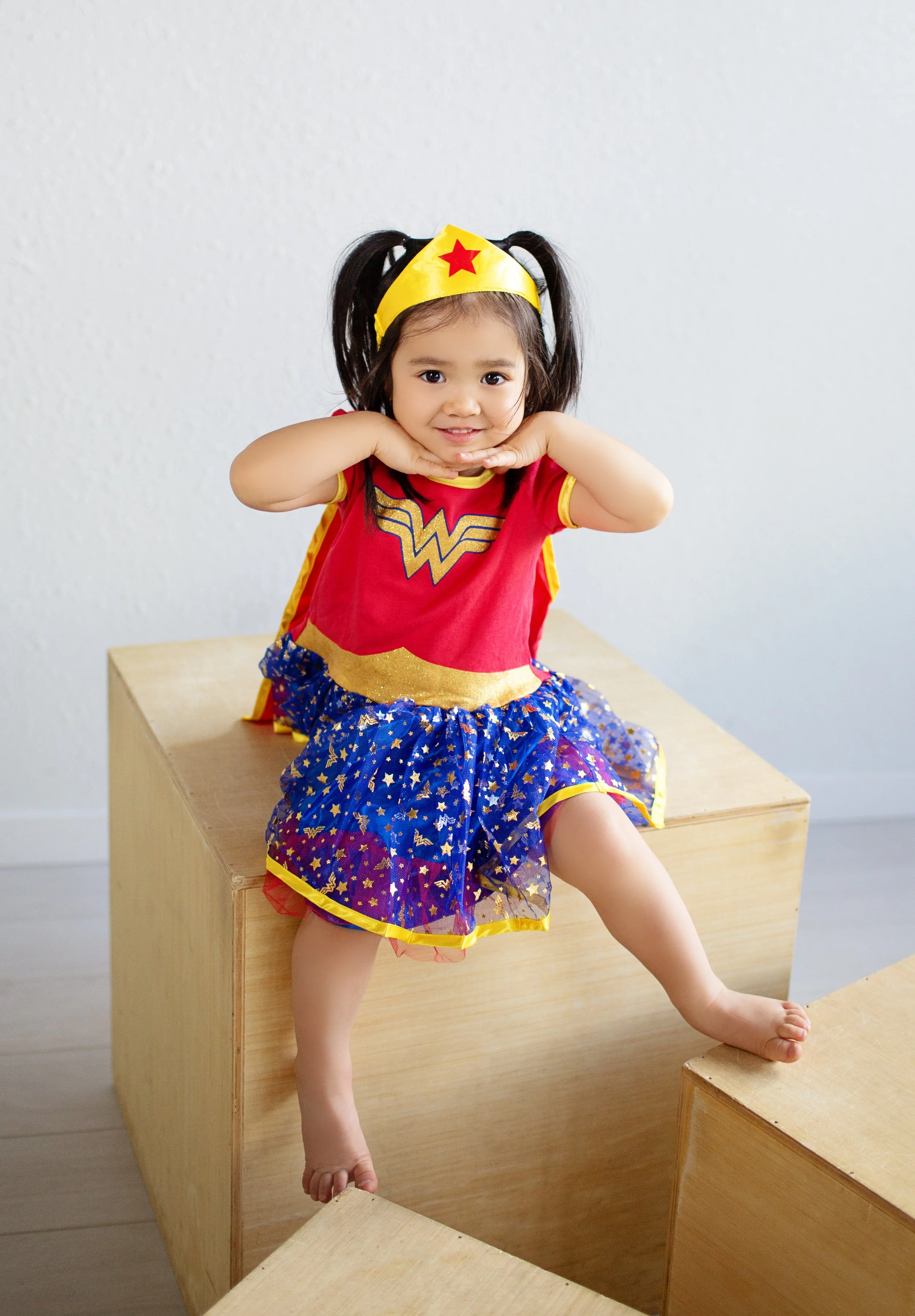 Young girl in Wonder Woman costume sitting on wooden blocks, making a playful pose.