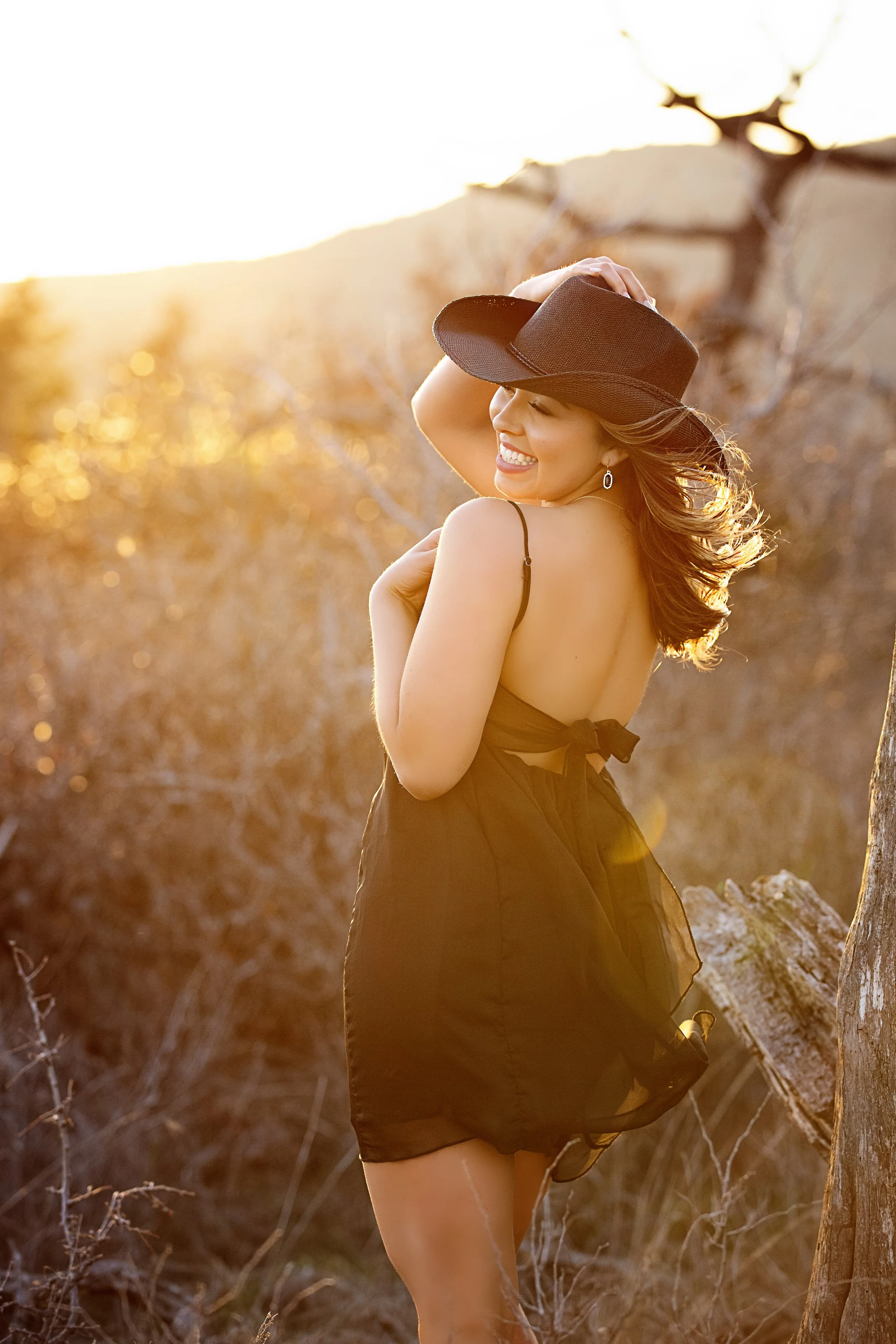 A woman smiling and wearing a black dress and a wide-brimmed hat, standing outdoors during sunset with trees and mountains in the background.