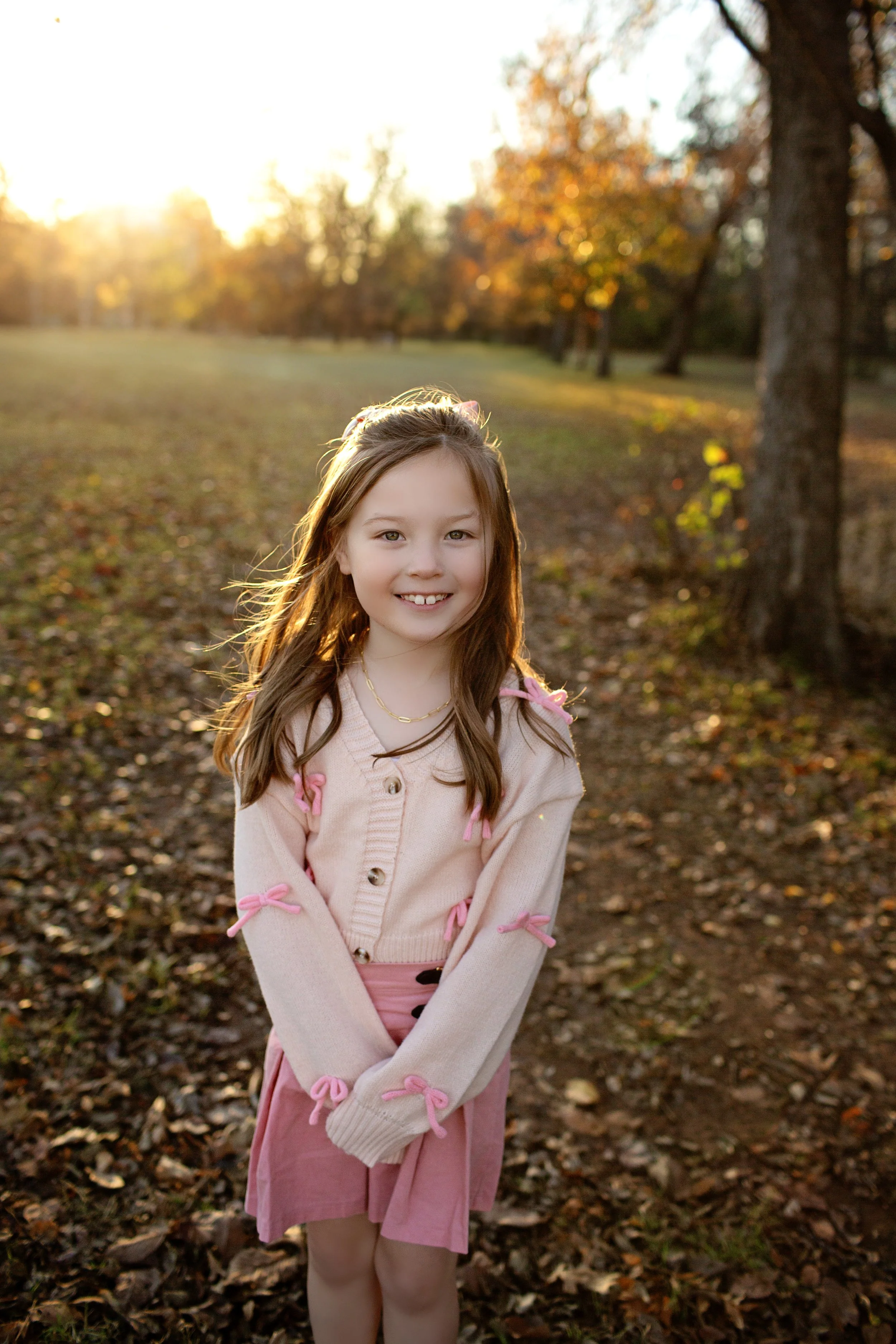 A young girl with long brown hair and a pink cardigan standing outdoors on a fall day, smiling with autumn trees and sunlight in the background.