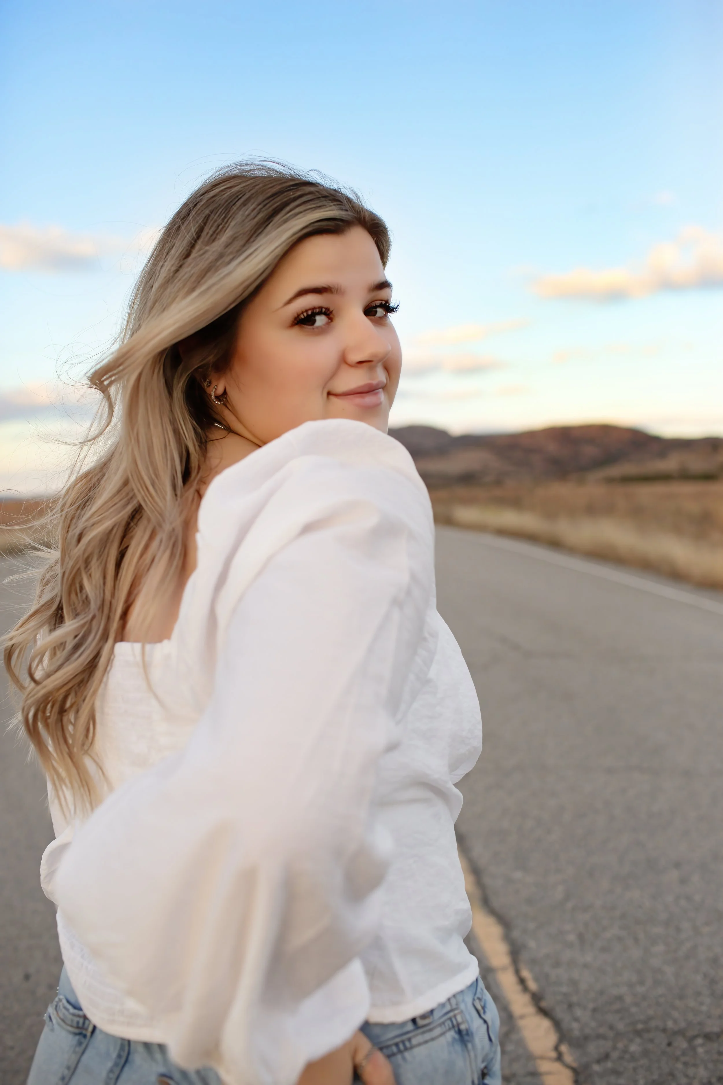 Young woman with wavy blonde hair smiling and looking over her shoulder on an open road during sunset.
