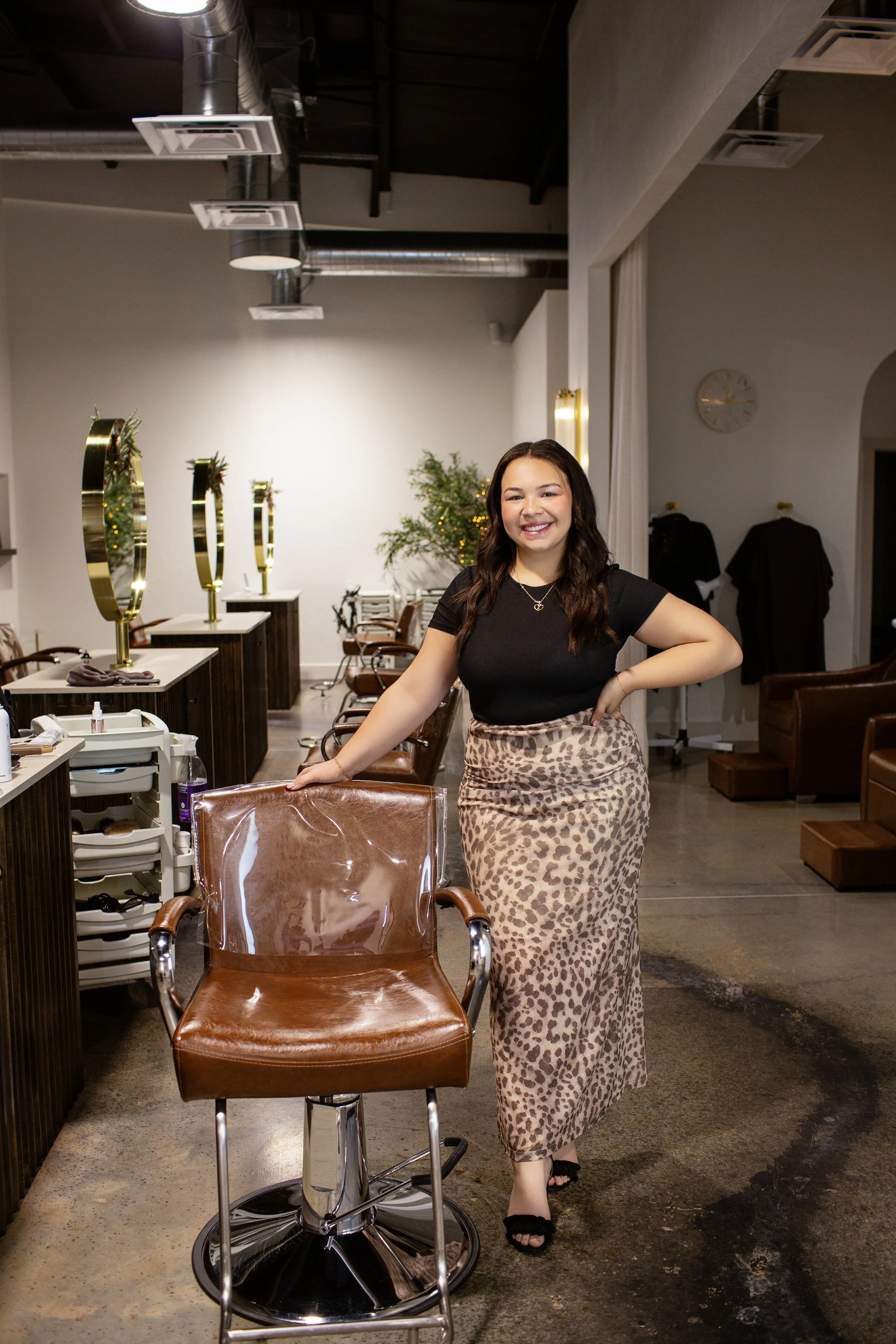 Smiling woman in black top and leopard print skirt in hair salon with styling chairs, mirrors, and salon equipment.
