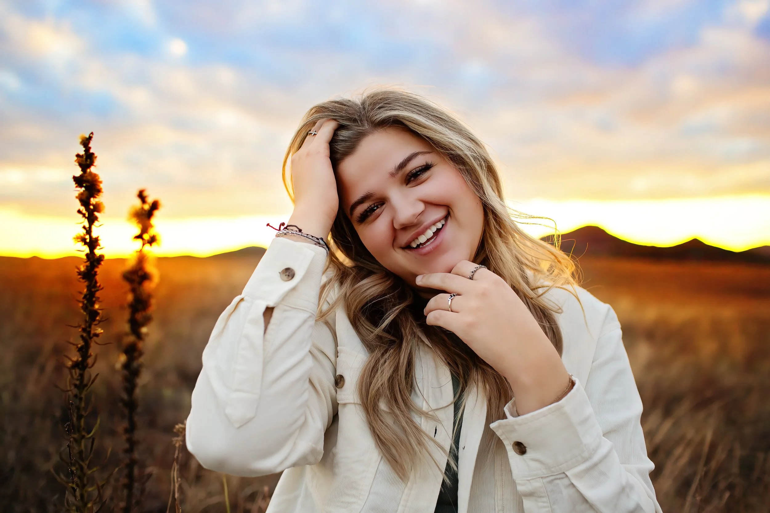 A young woman with blonde hair smiling outdoors during sunset, wearing a white jacket, standing in a field with tall grass and mountains in the background.