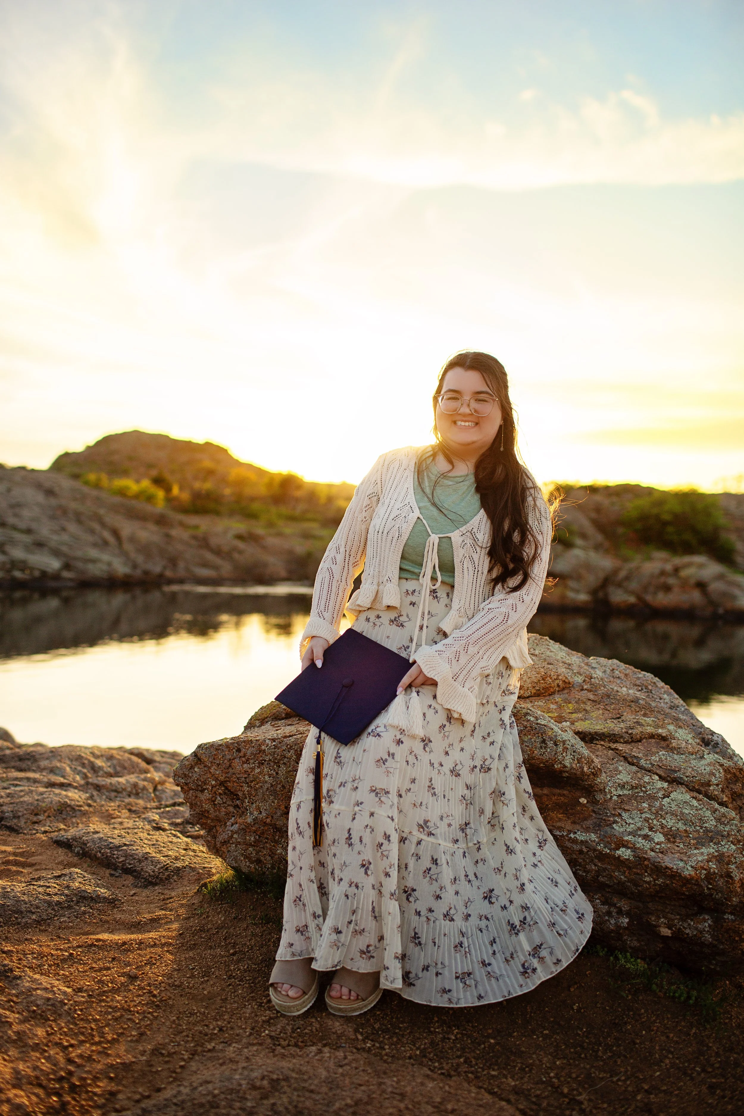 A young woman standing on a rocky terrain with a lake and hills in the background during sunset, holding a graduation cap and smiling.