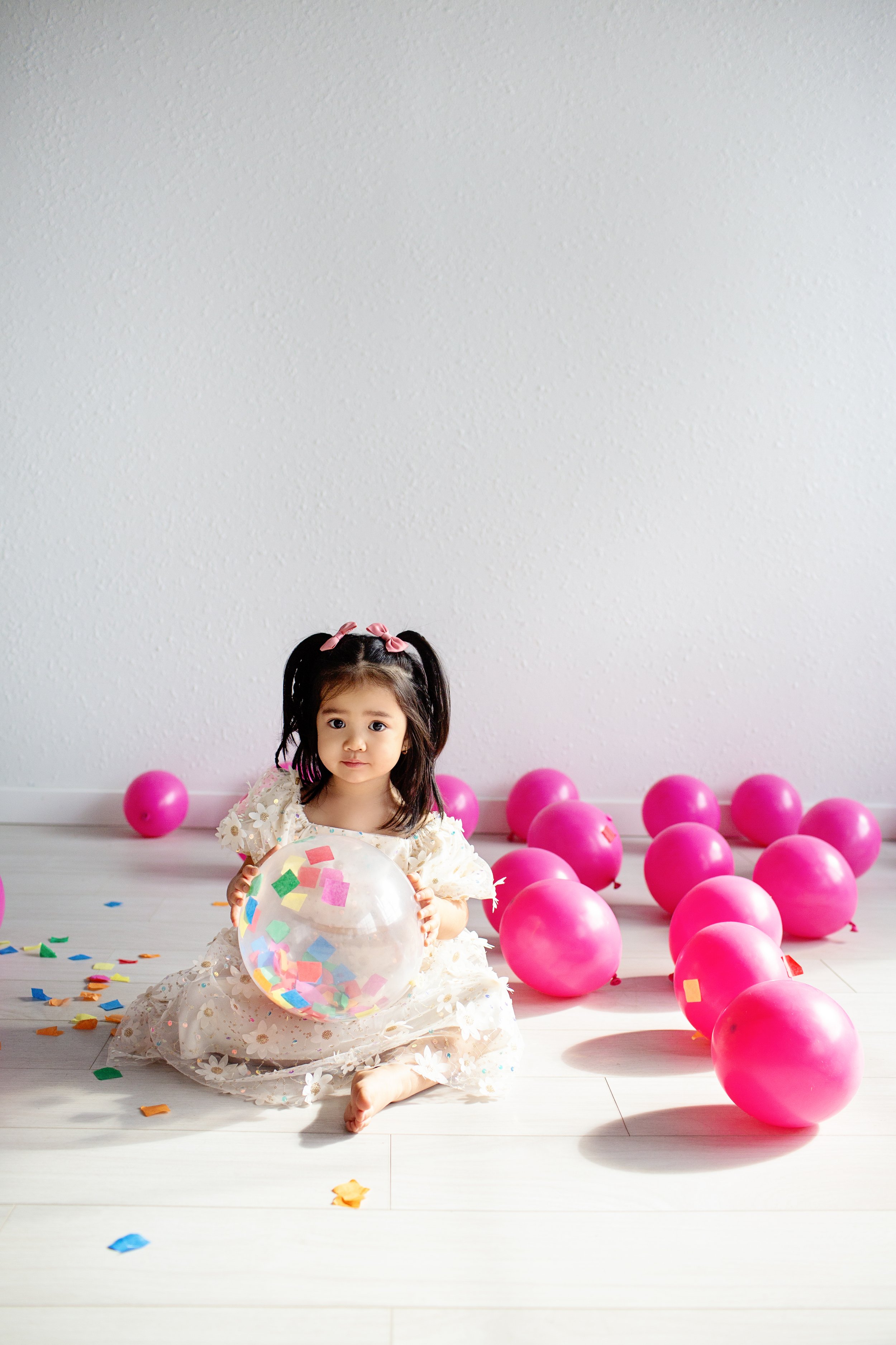 A young girl with pigtails and a floral dress sitting on the floor surrounded by pink balloons and colorful confetti, holding a transparent balloon filled with multicolored paper pieces.