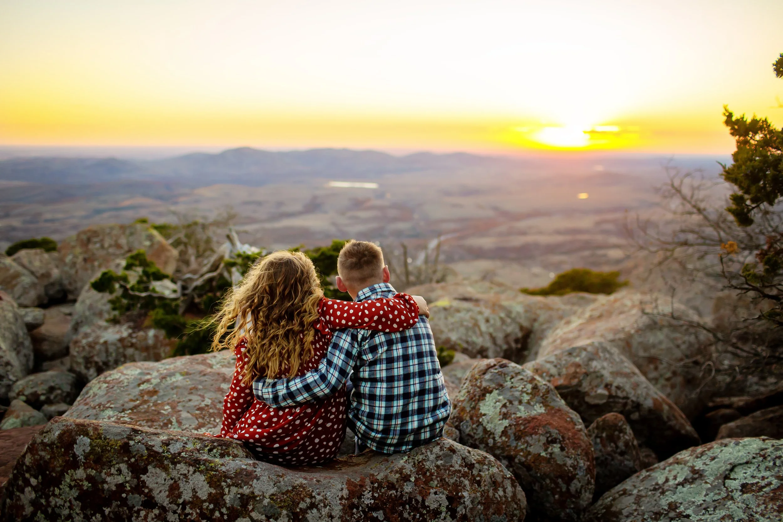 Two children, a girl with curly hair in a red dress with white polka dots and a boy with short hair in a blue plaid shirt, sit on rocks embracing as they watch the sunset over a mountainous landscape.