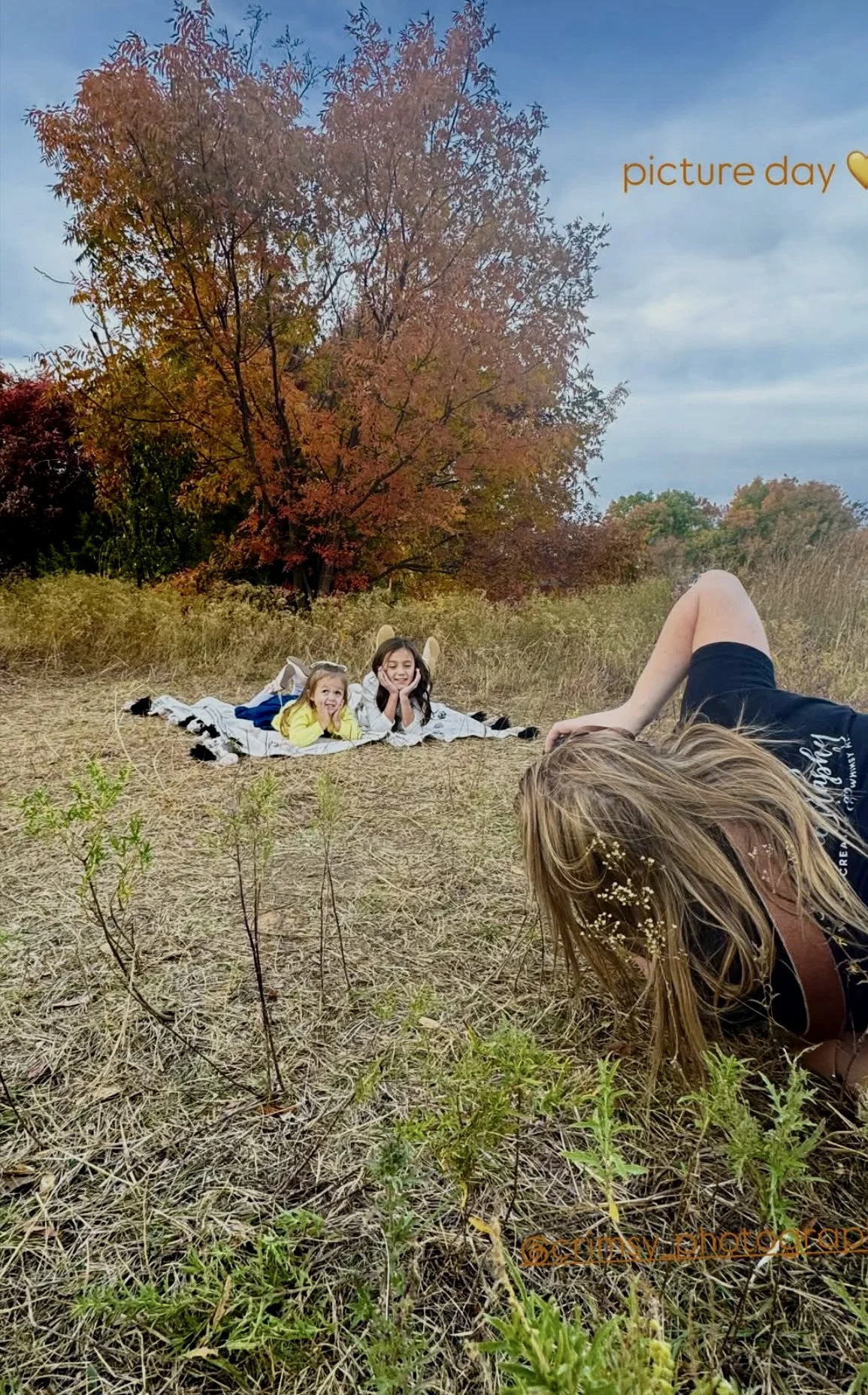 A woman lying on the ground taking a photo of two children lying on a blanket in an outdoor field with fall foliage in the background. The scene depicts a fall day, as indicated by the text 'picture day' with a yellow heart.