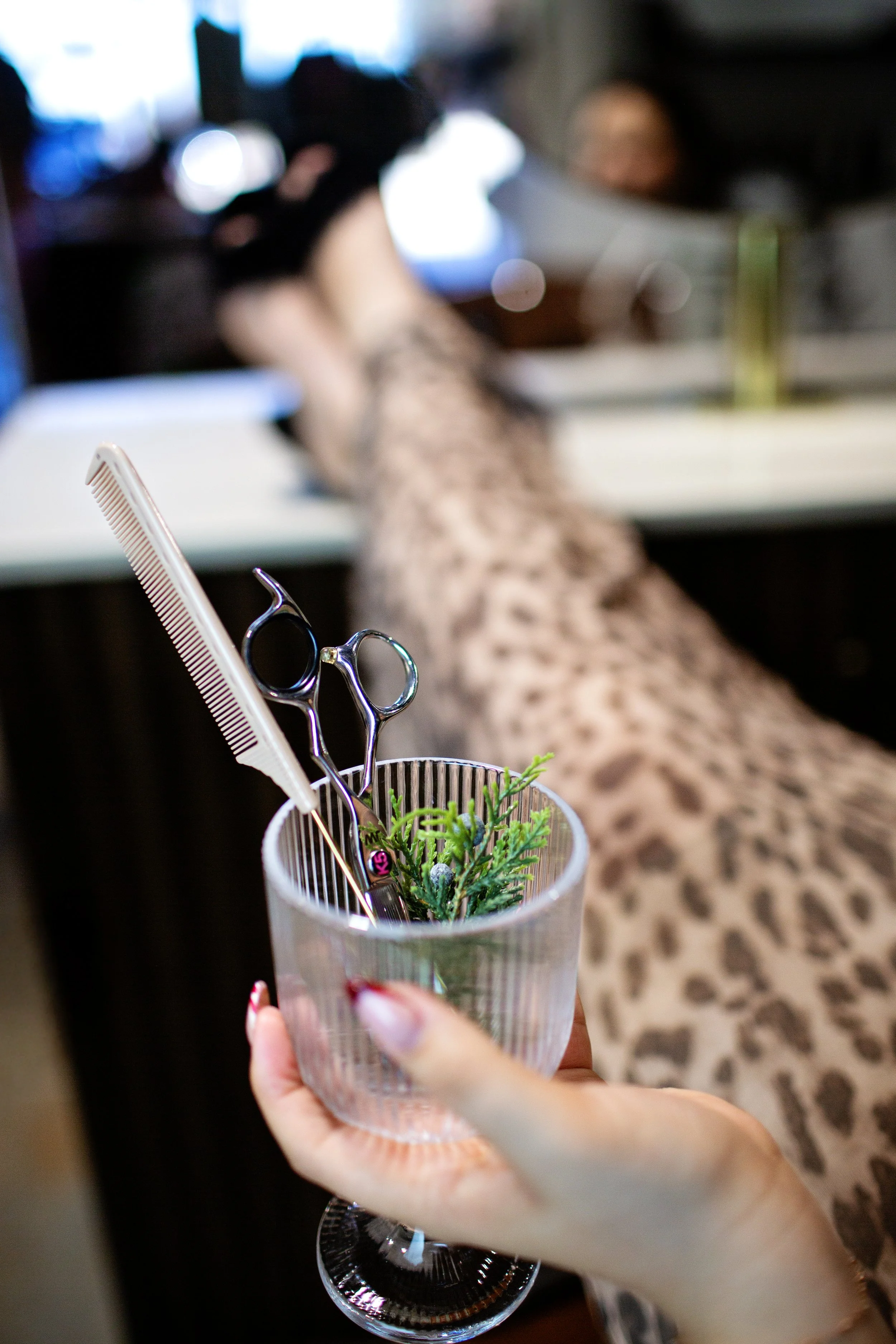 A person holding a glass with small pair of scissors, a comb, and faux green plant inside, with a woman getting a haircut in the background.
