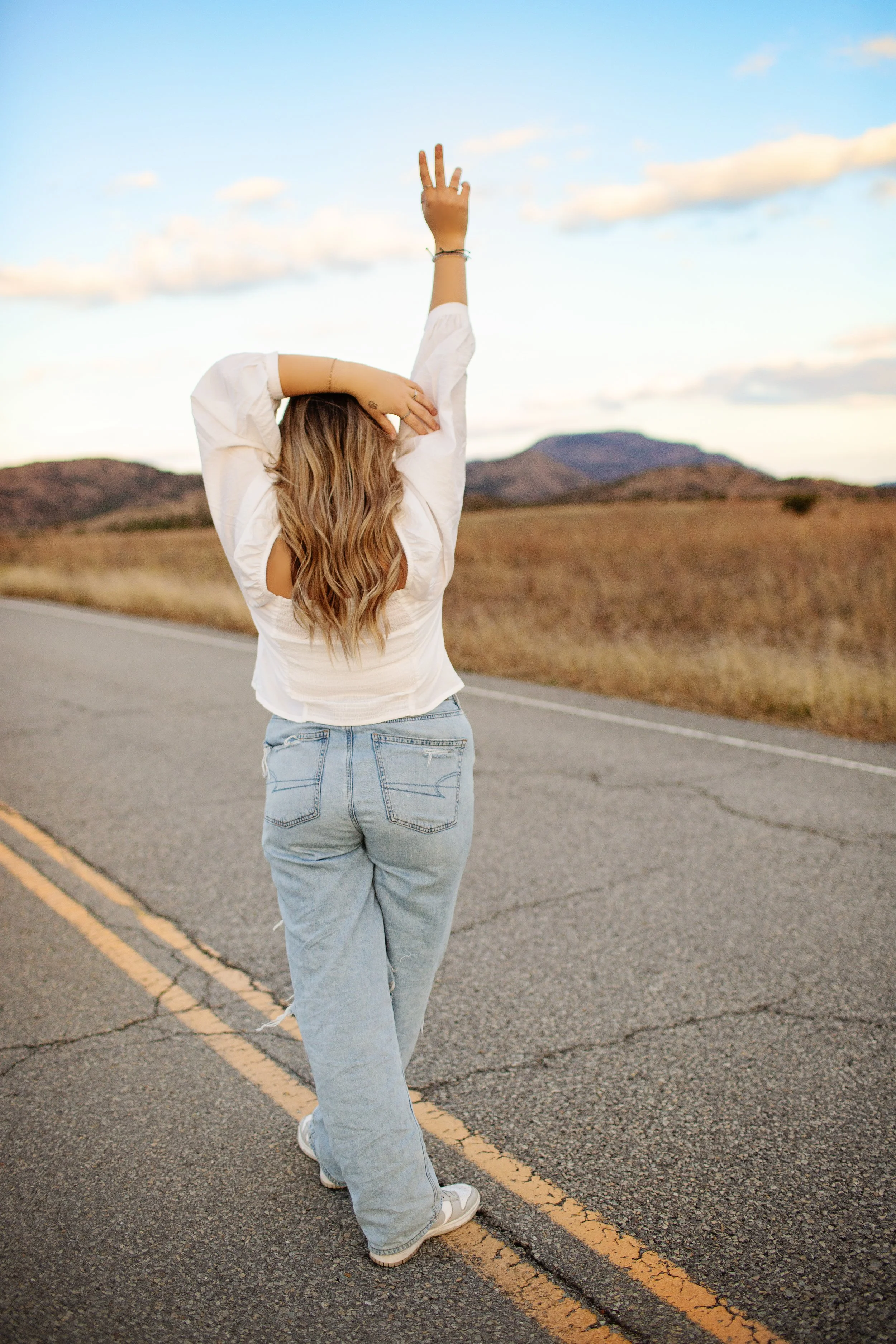 A woman with long, wavy hair stands on an empty rural road, stretching her right arm upward in a peaceful landscape with hills in the background during sunset.