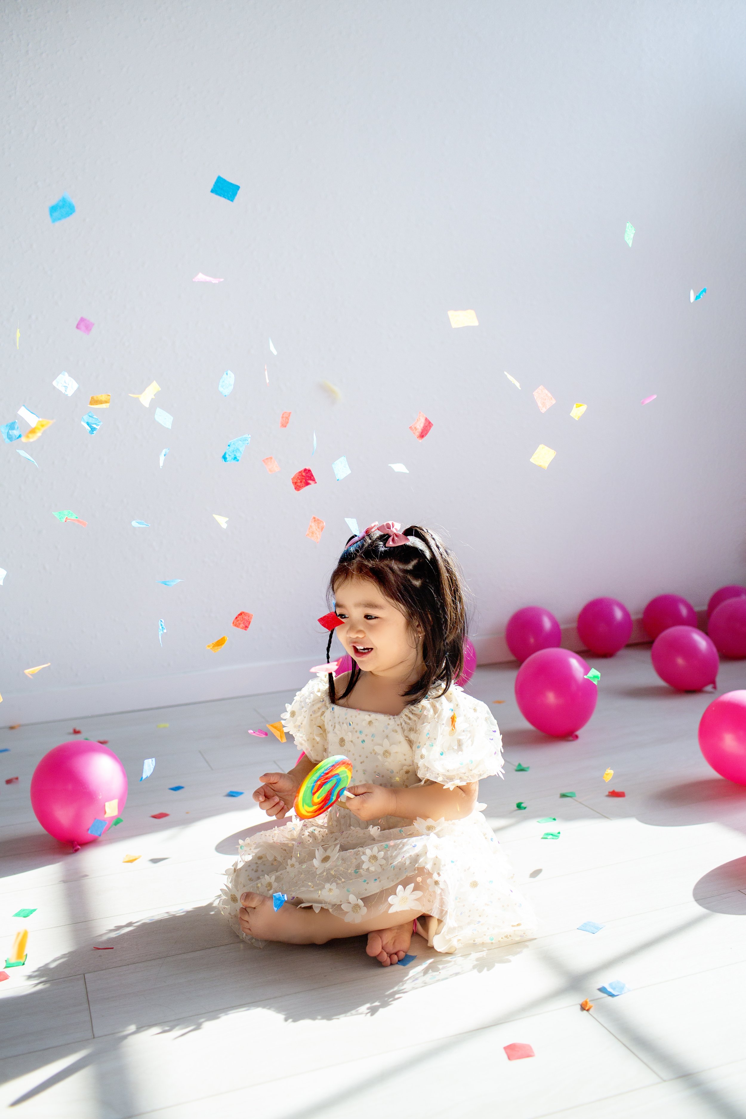 A young girl sitting on the floor holding a colorful lollipop during a birthday celebration with pink balloons and confetti around her.