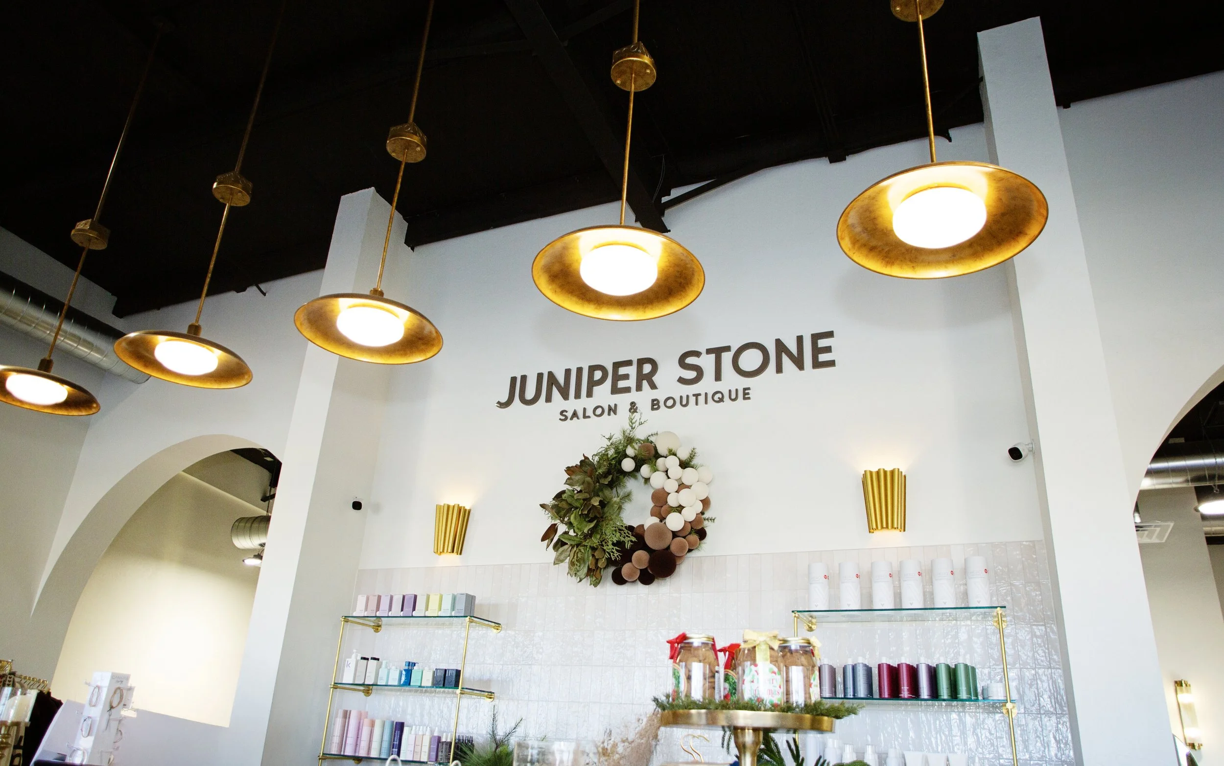 Interior view of Juniper Stone Salon & Boutique, featuring hanging gold lights, a decorative wreath on the wall, and shelves with products.