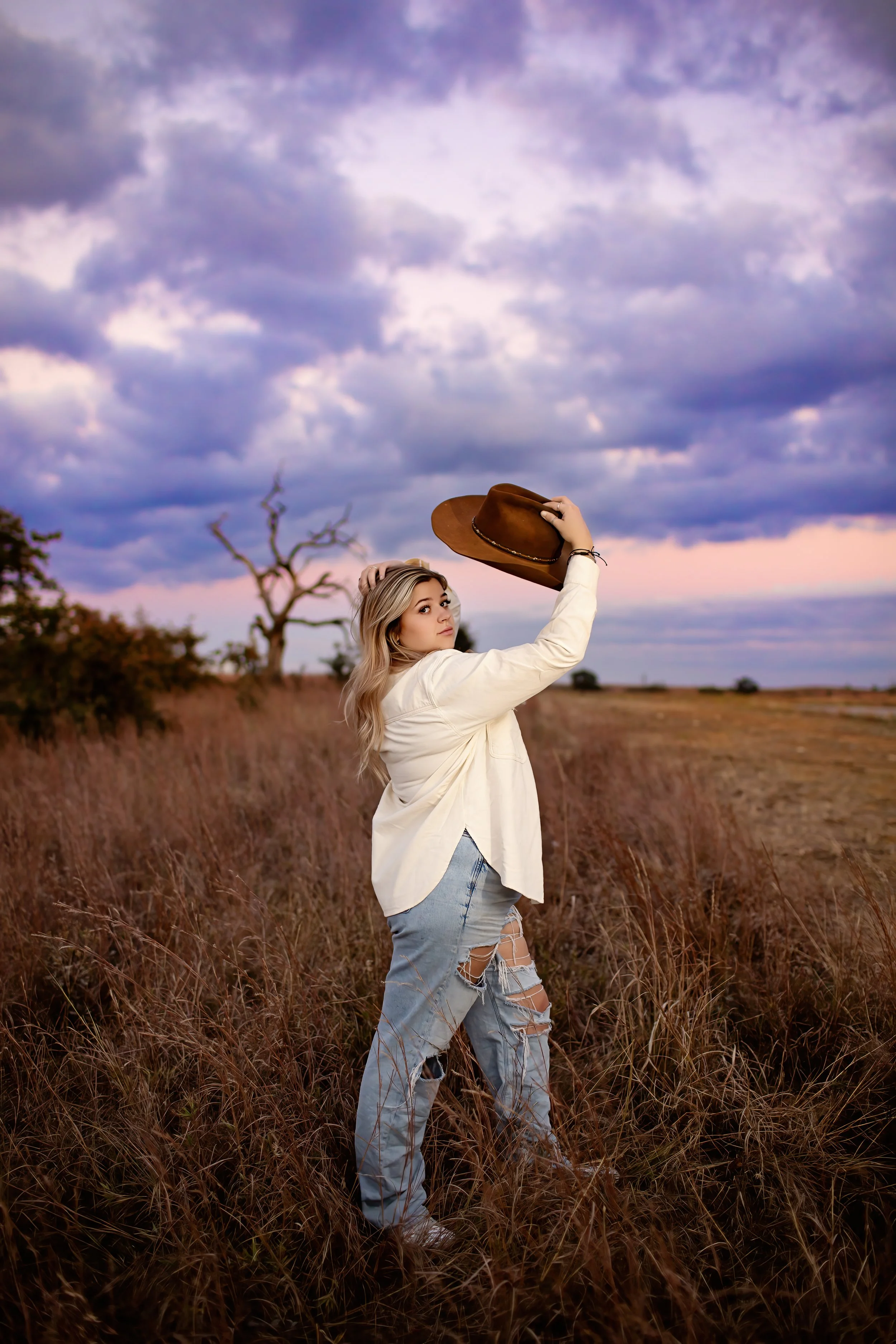 A young woman with blonde hair in a field holding a brown cowboy hat, wearing a white shirt and ripped jeans, with a sunset sky and clouds in the background.