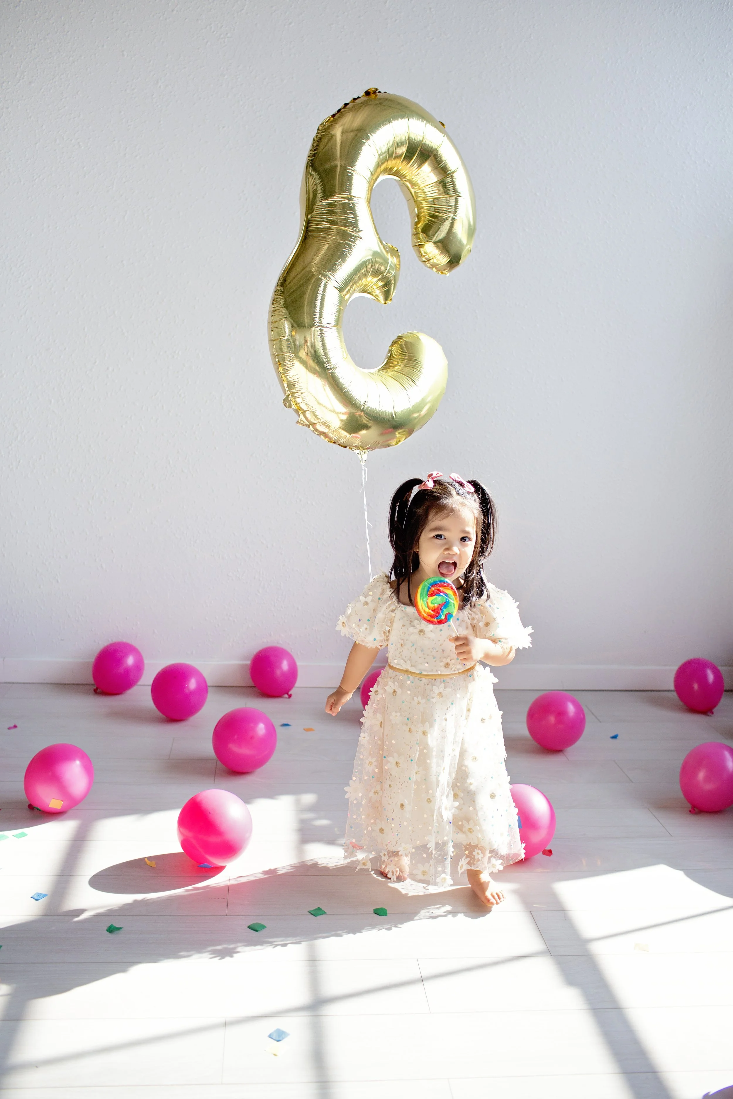 A young girl at a birthday celebration holding a colorful lollipop and standing on a wooden floor, surrounded by pink balloons and a large gold foil balloon shaped like the number '2'.