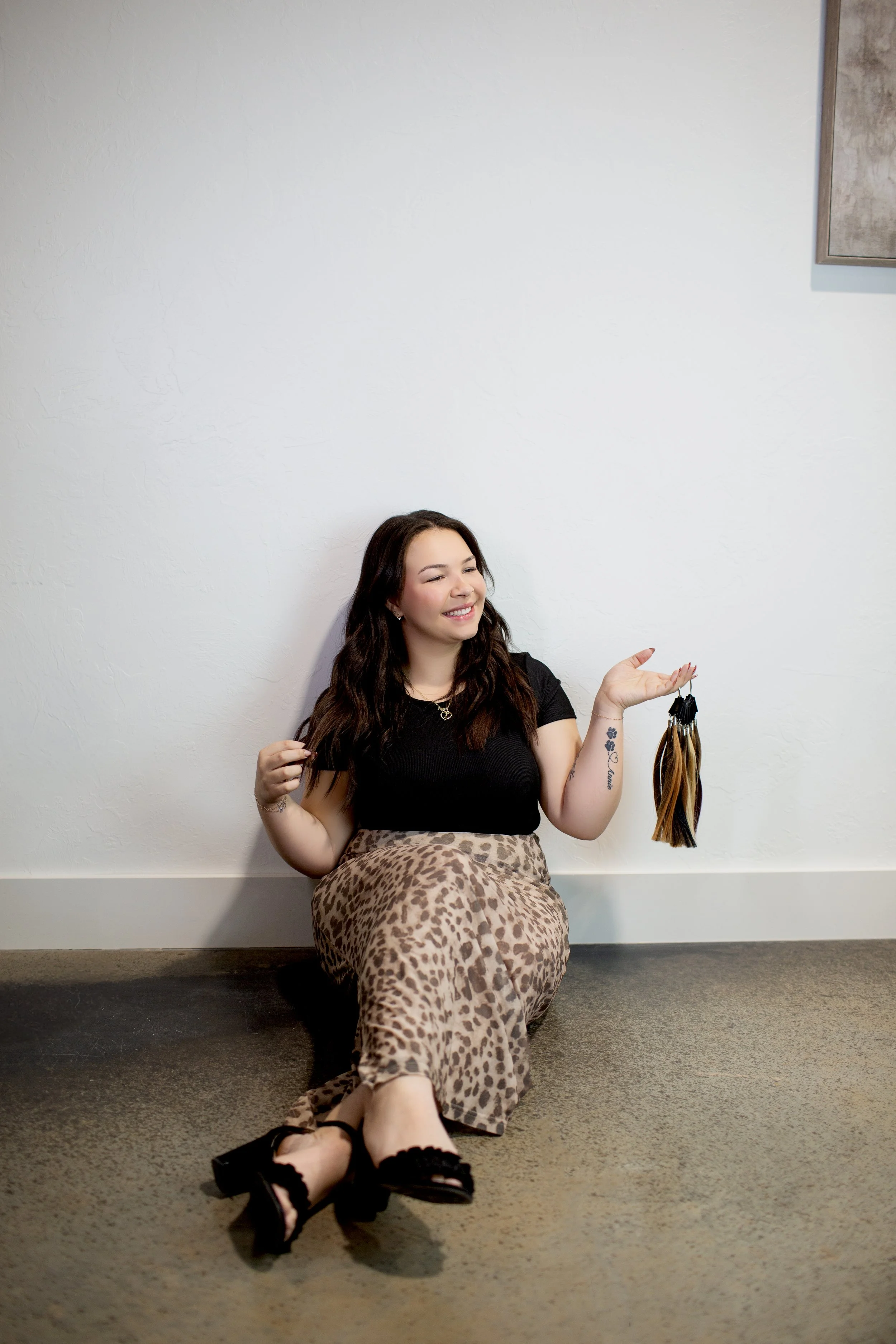 Woman sitting on the floor with her back against a wall, holding a sample of hair extensions, smiling with eyes closed, wearing a black top, leopard print skirt, and black heels.