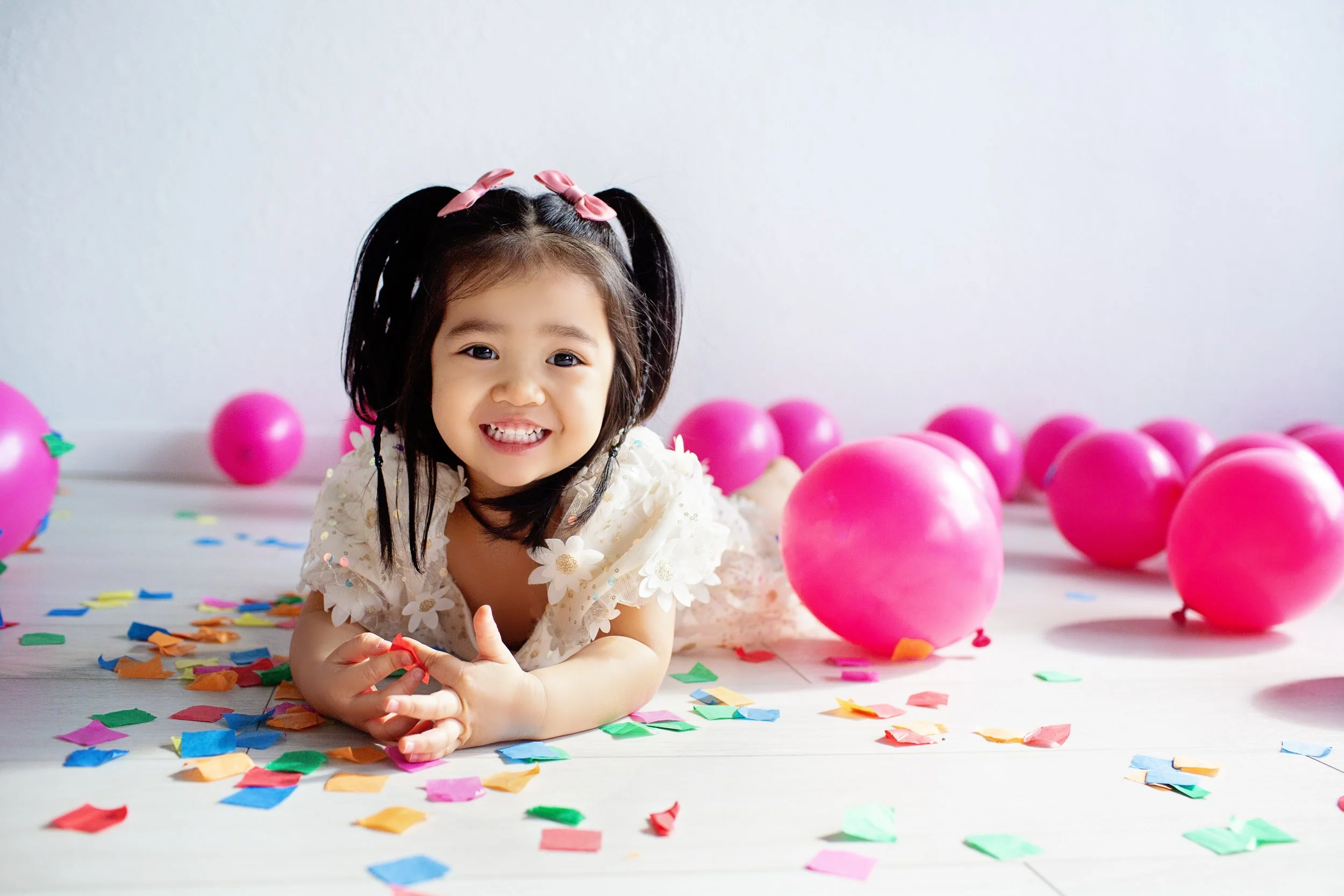 A young girl with black hair and pink bows, smiling and lying on the floor surrounded by pink balloons and colorful confetti.
