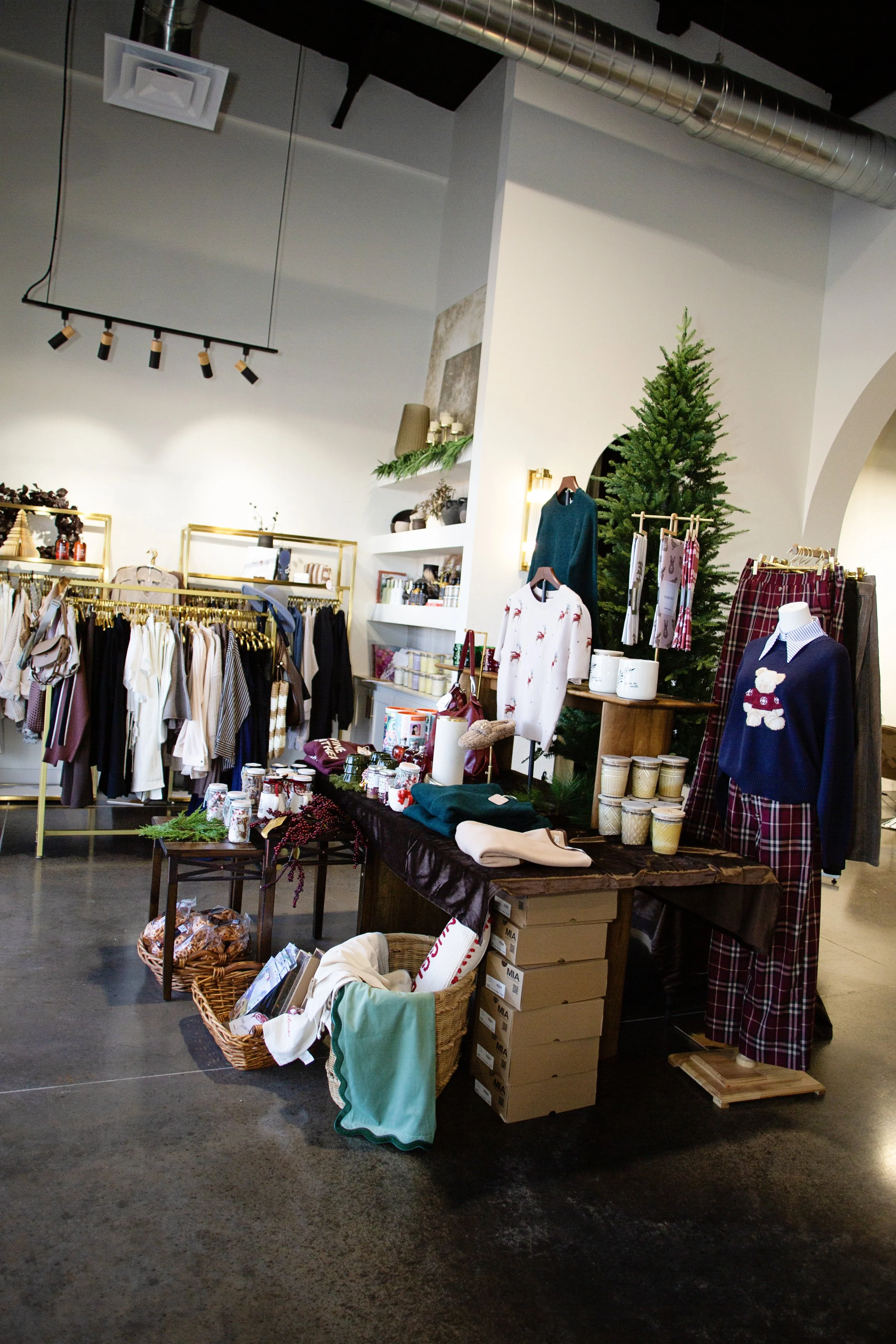 Holiday-themed clothing and accessories display in a retail store with a Christmas tree, sweaters, pajamas, and gift items.