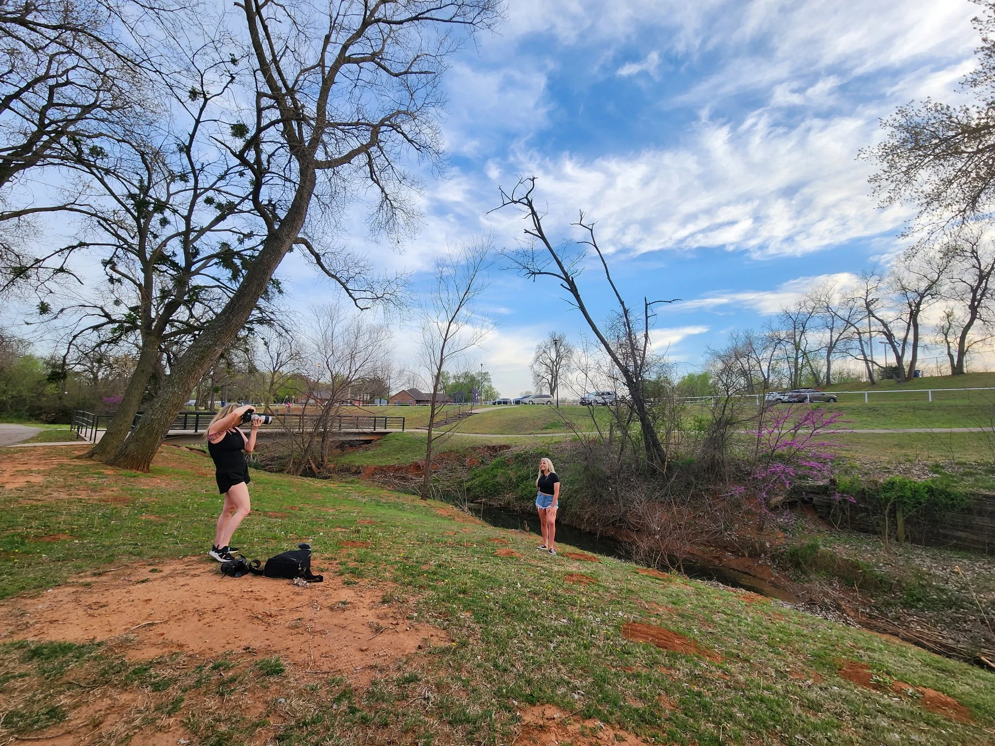 A woman taking a photo of another woman standing near a body of water with trees and a grassy area in the background.