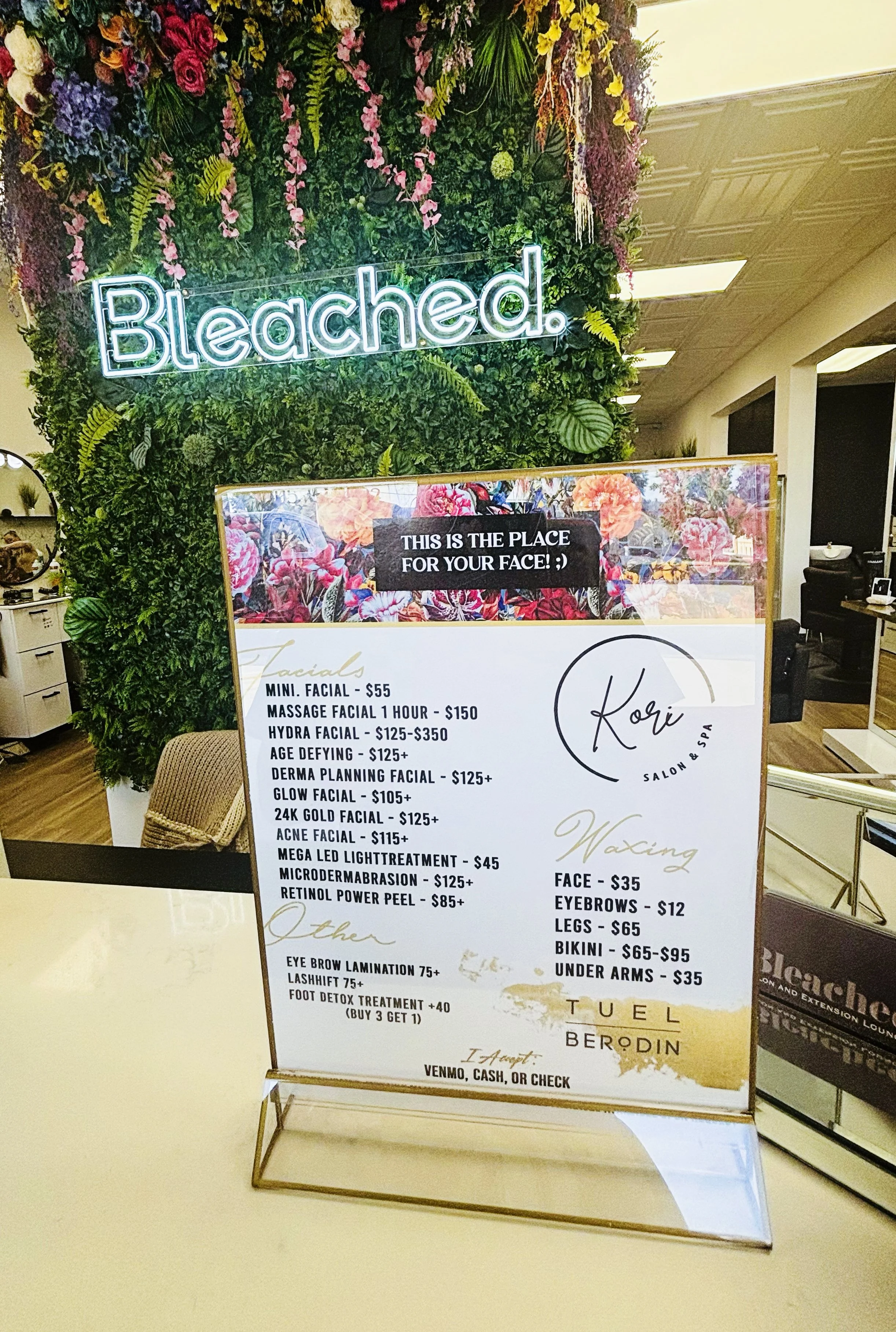 A flower wall with a neon sign that says 'Bleached.' In front of the wall, there is a menu board listing facial and waxing services at a spa, with various prices. The surrounding decor includes a workspace area in the background.