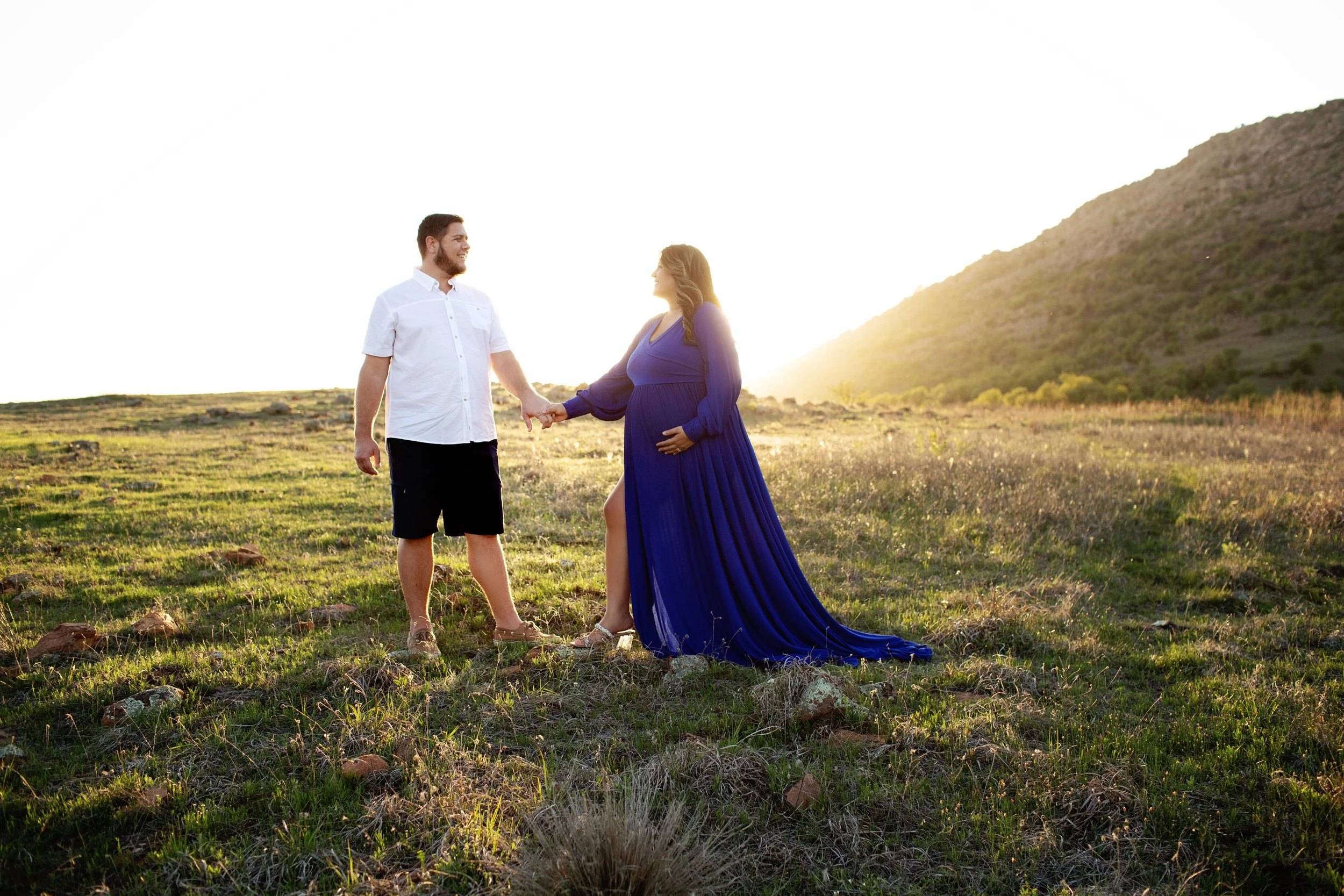 A pregnant woman in a blue gown holding hands with a man in casual attire in a grassy field at sunset.