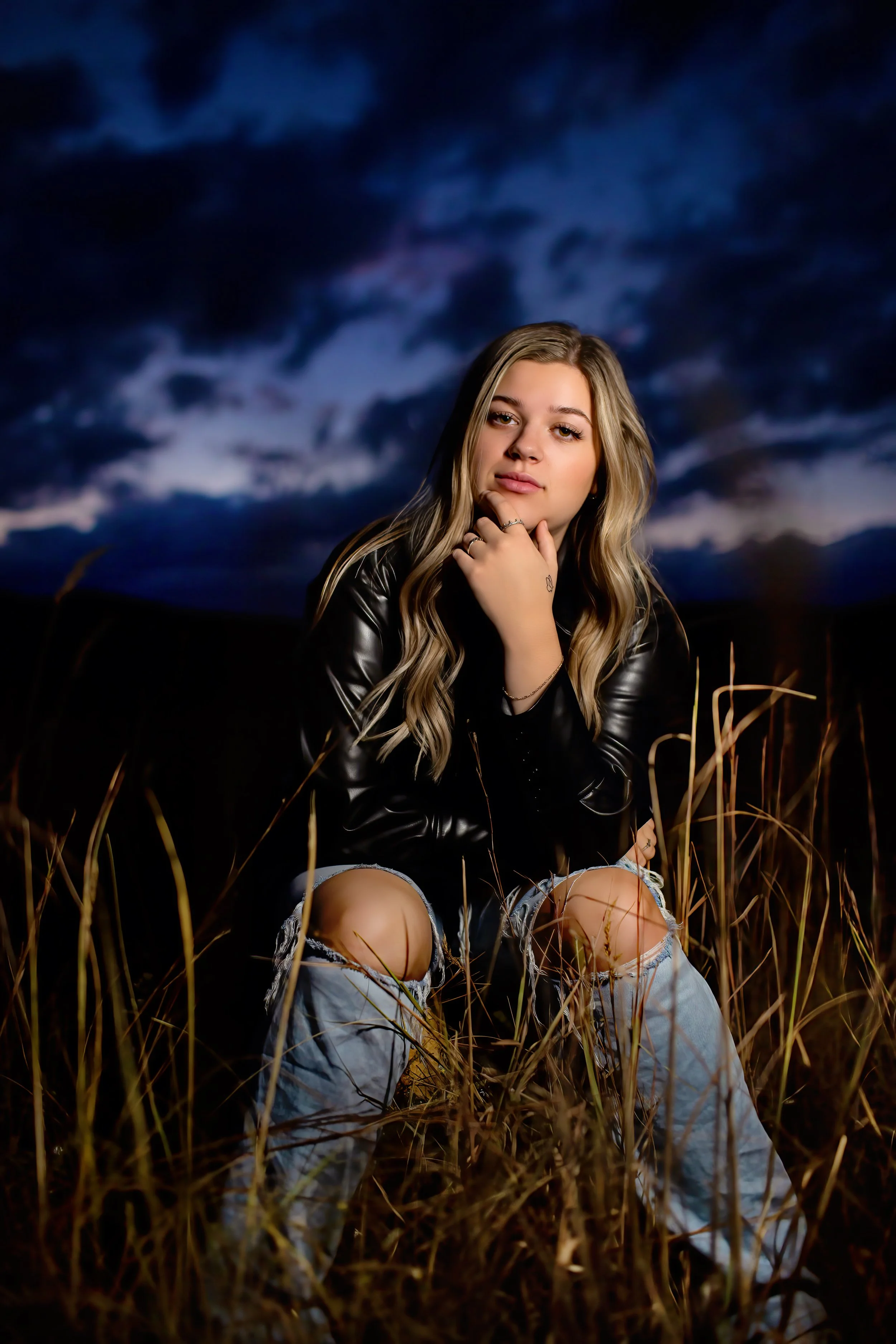 A young woman with blonde hair, wearing a black leather jacket and ripped jeans, sitting in a field of tall grass during dusk with a dark, cloudy sky in the background.