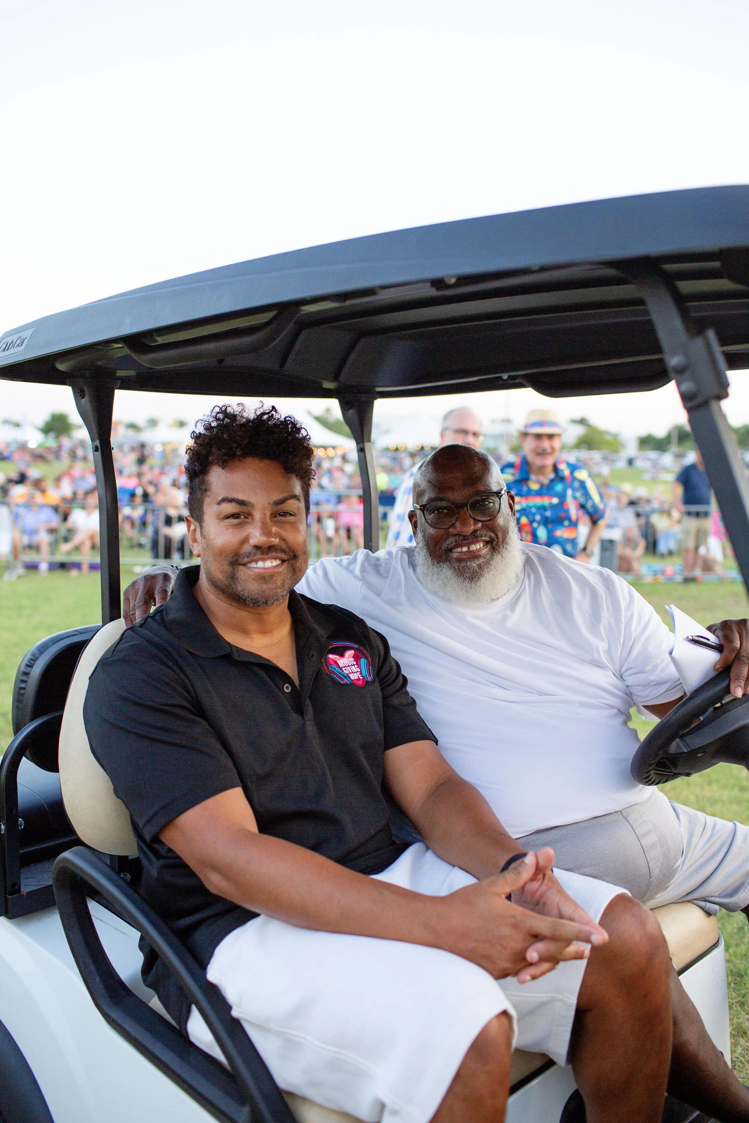 Two men sitting in a golf cart at an outdoor event, smiling, with a crowd in the background.