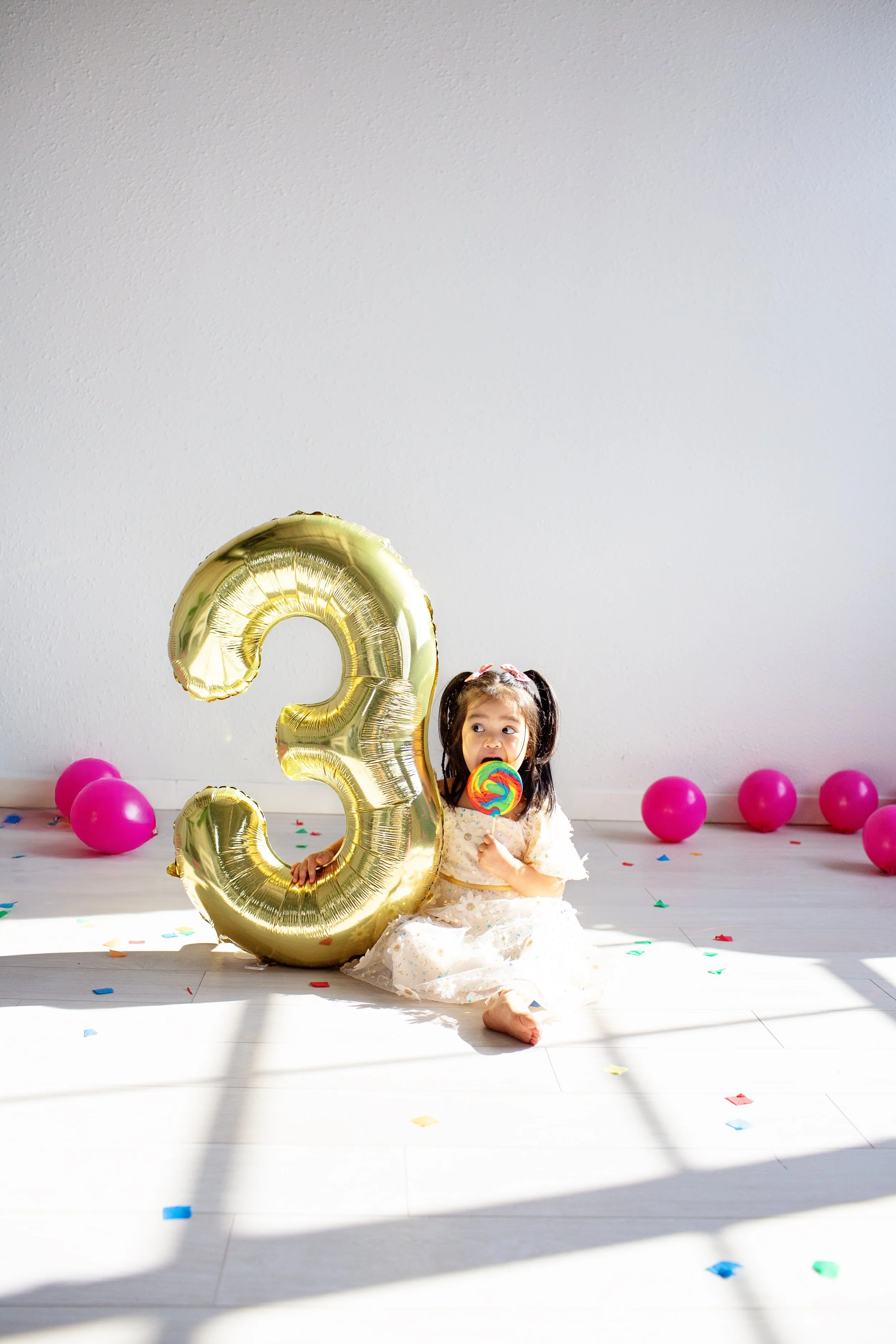 A young girl sits on the floor holding a large gold '3' balloon and a colorful lollipop in a celebration setting, with pink balloons and confetti scattered around.