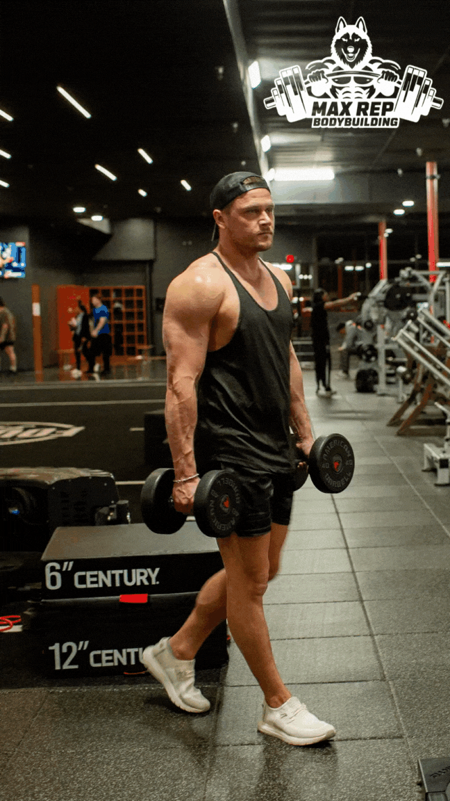 A muscular man in a black tank top and shorts, holding dumbbells, standing in a gym with fitness equipment around him.