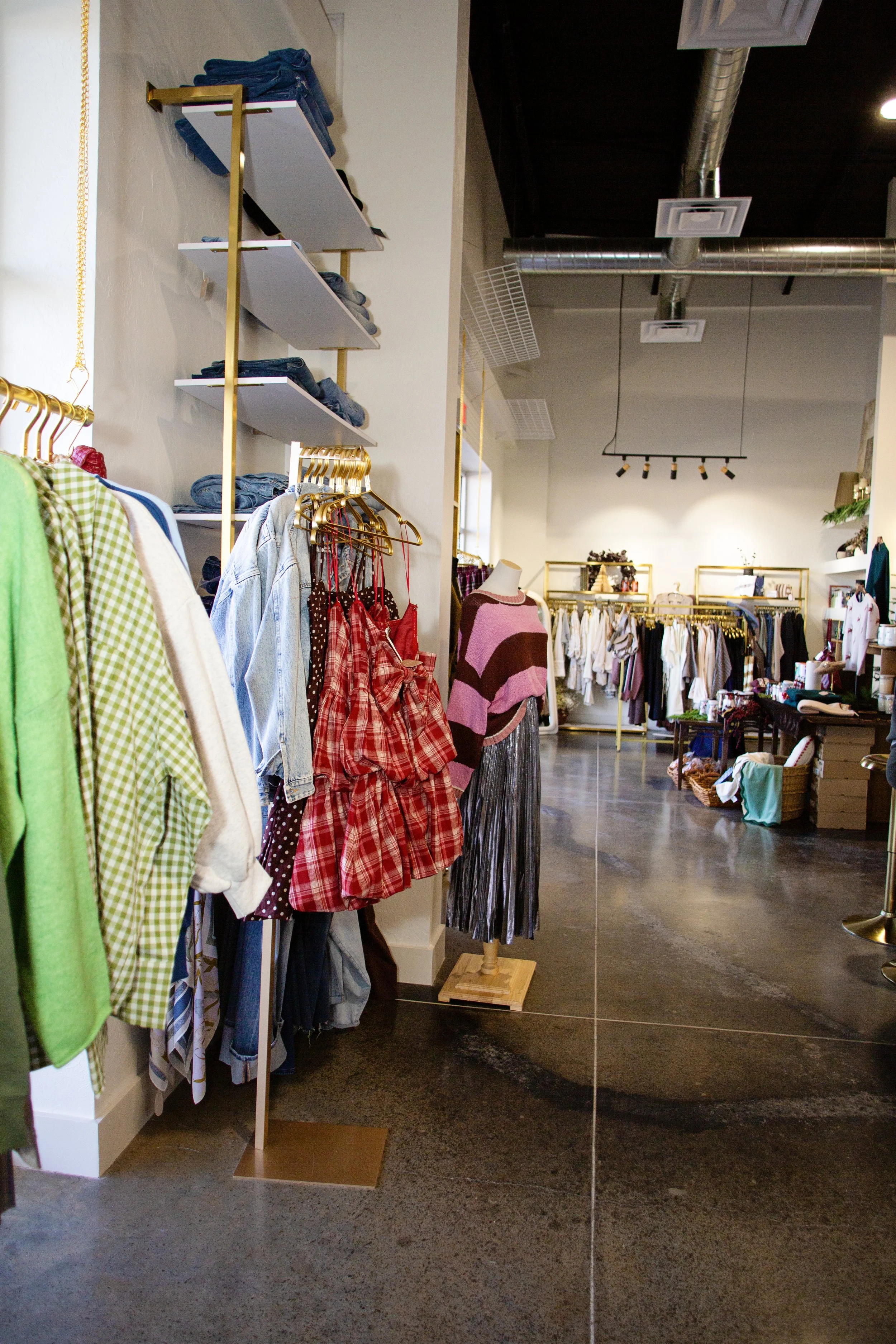 Clothing store with racks of clothes, including green, white, denim, red plaid dresses, a pink and brown sweater, and a metallic skirt on a mannequin. Shelves with folded jeans and miscellaneous items, and tables with more clothes and accessories in 