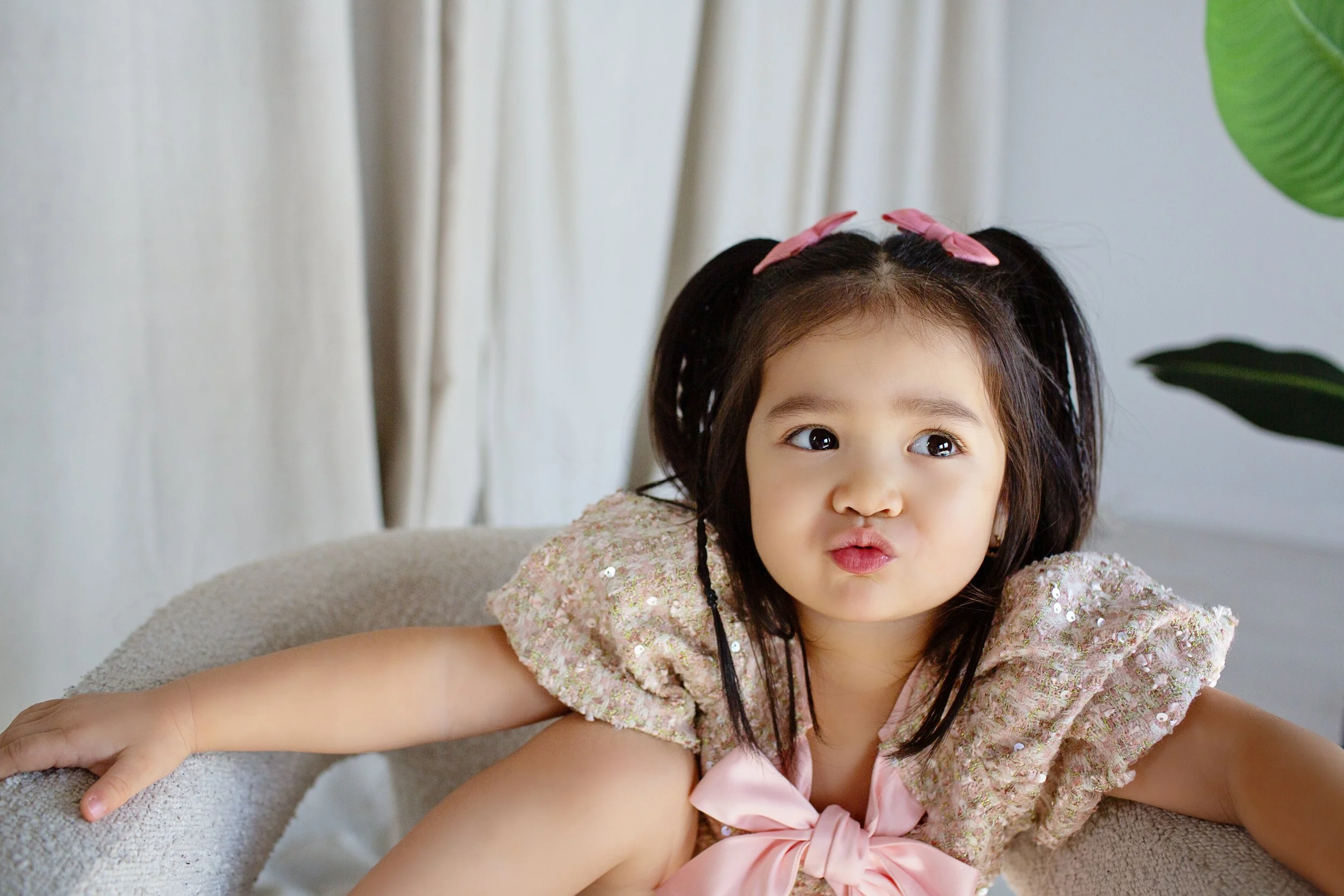 A young girl with pigtails and pink bows, wearing a sparkly pink top with a bow, making a playful face with puckered lips, sitting on a beige chair in a well-lit room with curtains and a green plant in the background.