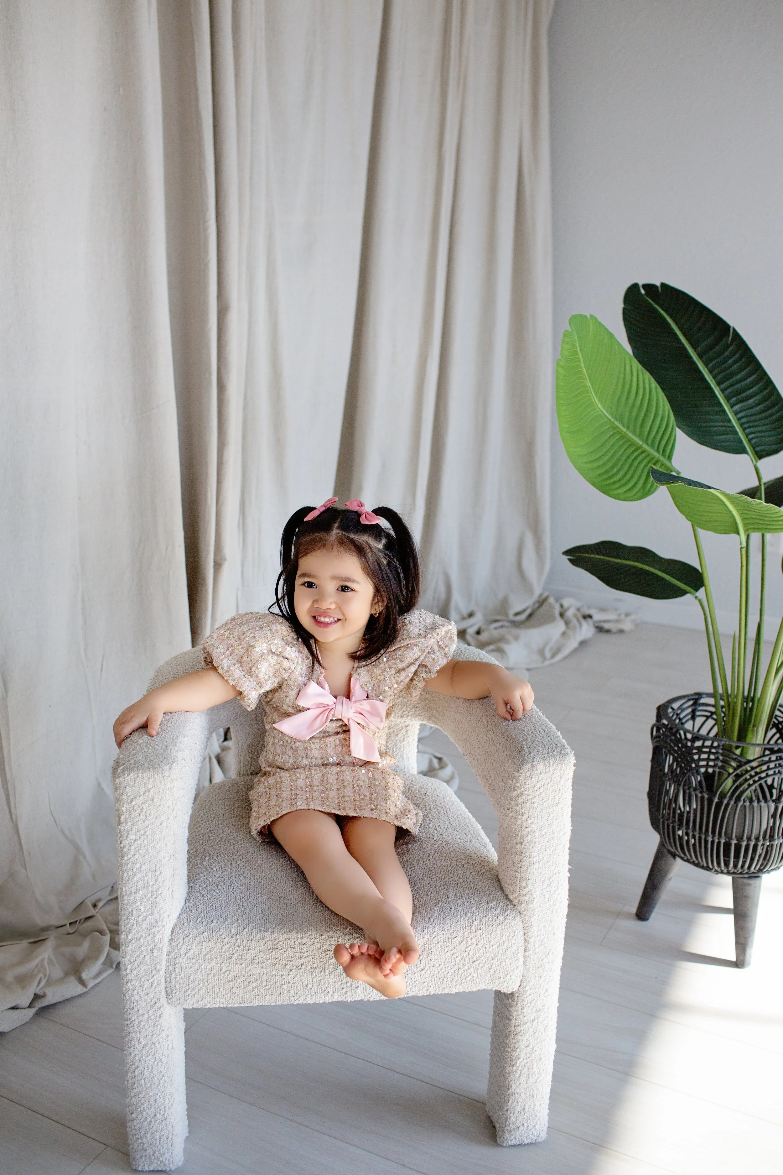 A young girl with dark hair in pigtails with pink bows, sitting on a white textured chair, smiling, wearing a pink and beige dress with puffed sleeves and a pink bow on the front, in a bright room with white curtains and a large potted green plant.