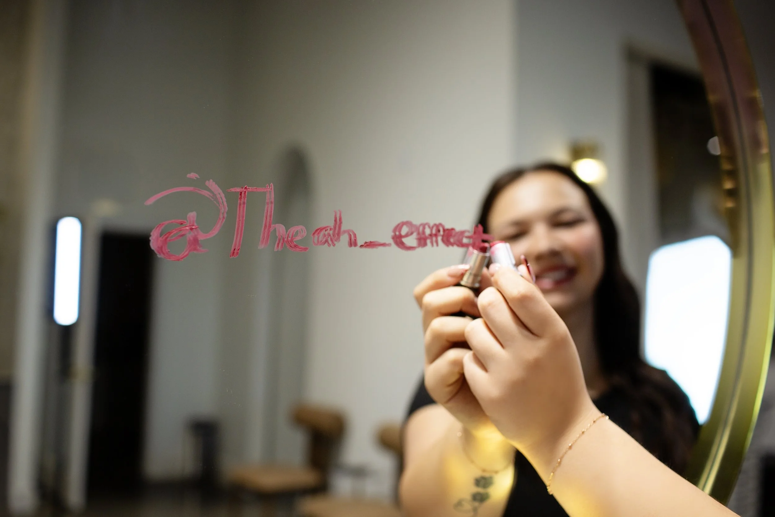 A woman writing on a mirror with pink lipstick, with a smile on her face, in a well-lit room.