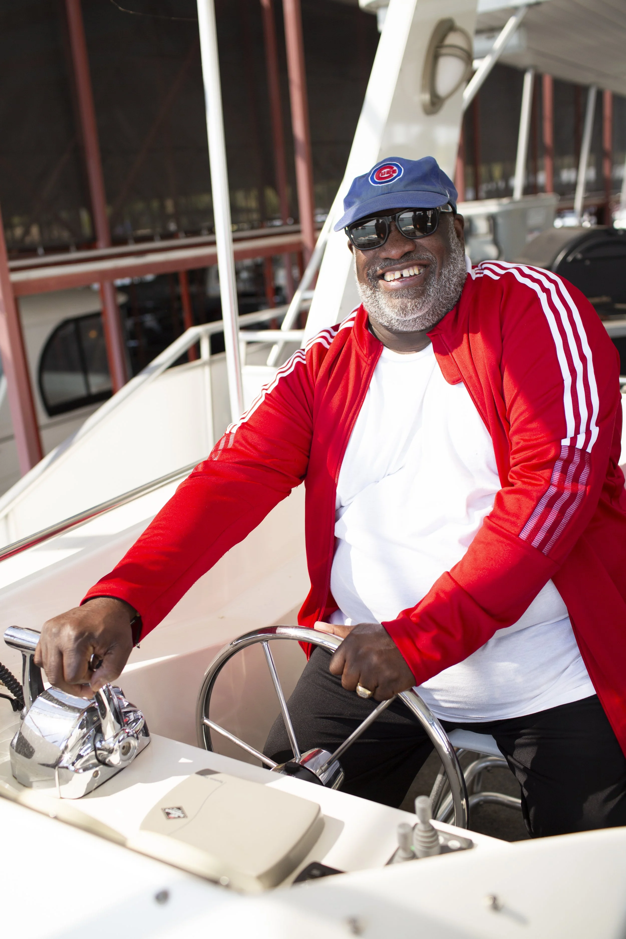 A smiling man with gray beard wearing sunglasses, a blue Chicago Cubs cap, a red jacket with white stripes, and a white T-shirt, sitting at the wheel of a boat.