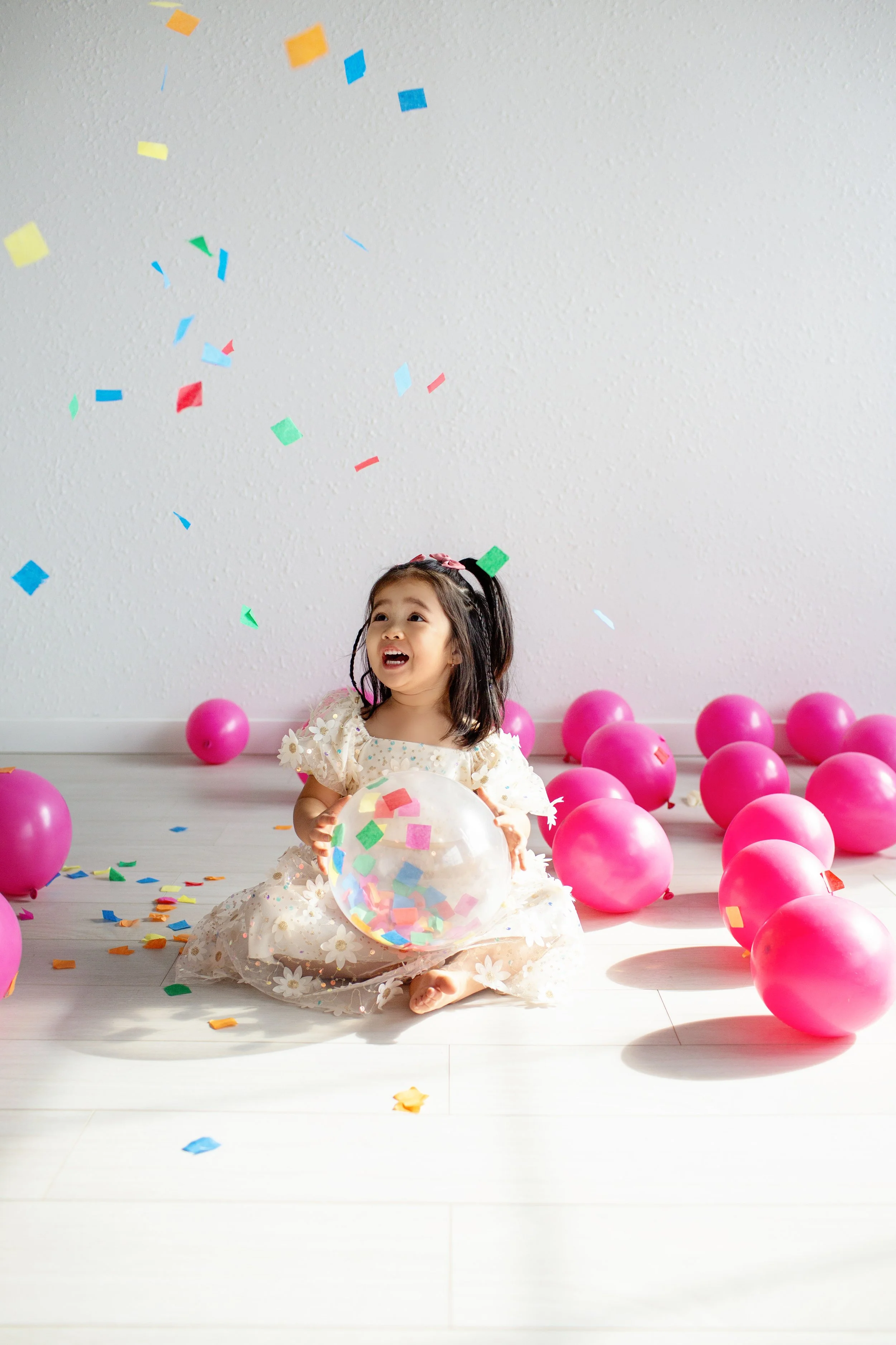 A young girl in a dress sitting on the floor, surrounded by pink balloons and colorful confetti, holding a clear ball with confetti inside, celebrating a party.